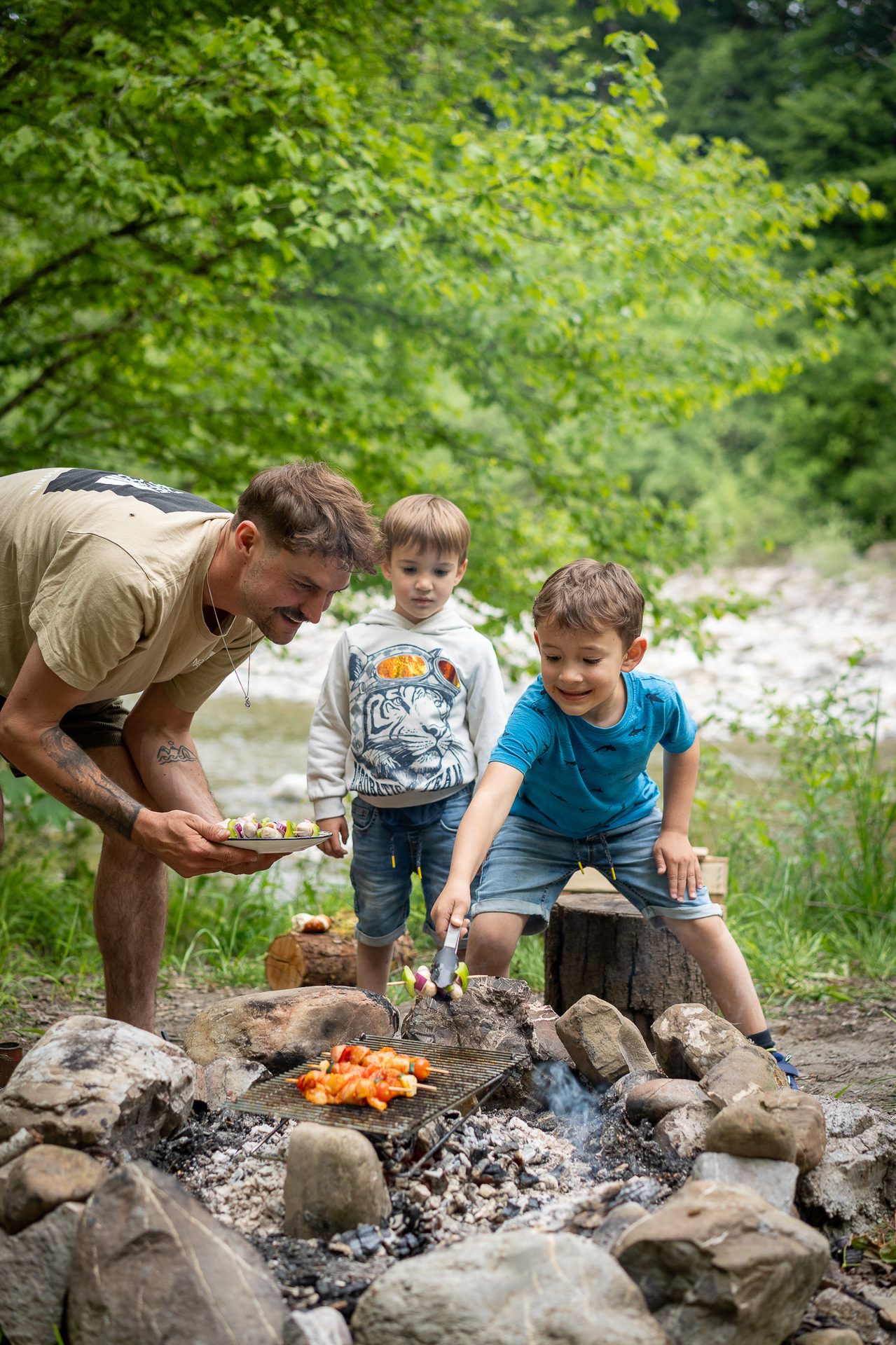 Father and two boys grilling skewers over a campfire in a lush green outdoor setting by a river.