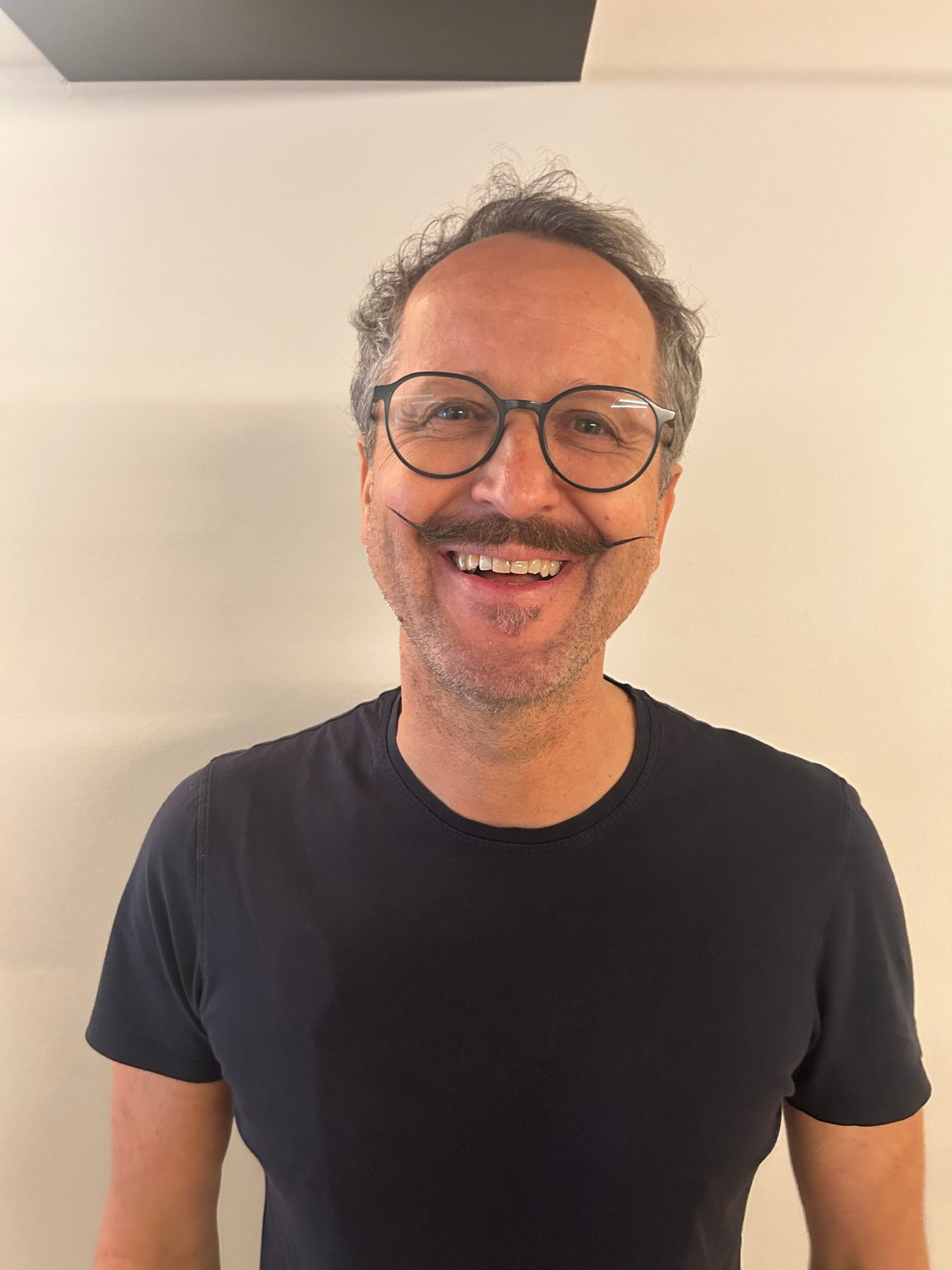 A smiling man with curly grey hair, round glasses, and a long, curly mustache wearing a dark t-shirt.