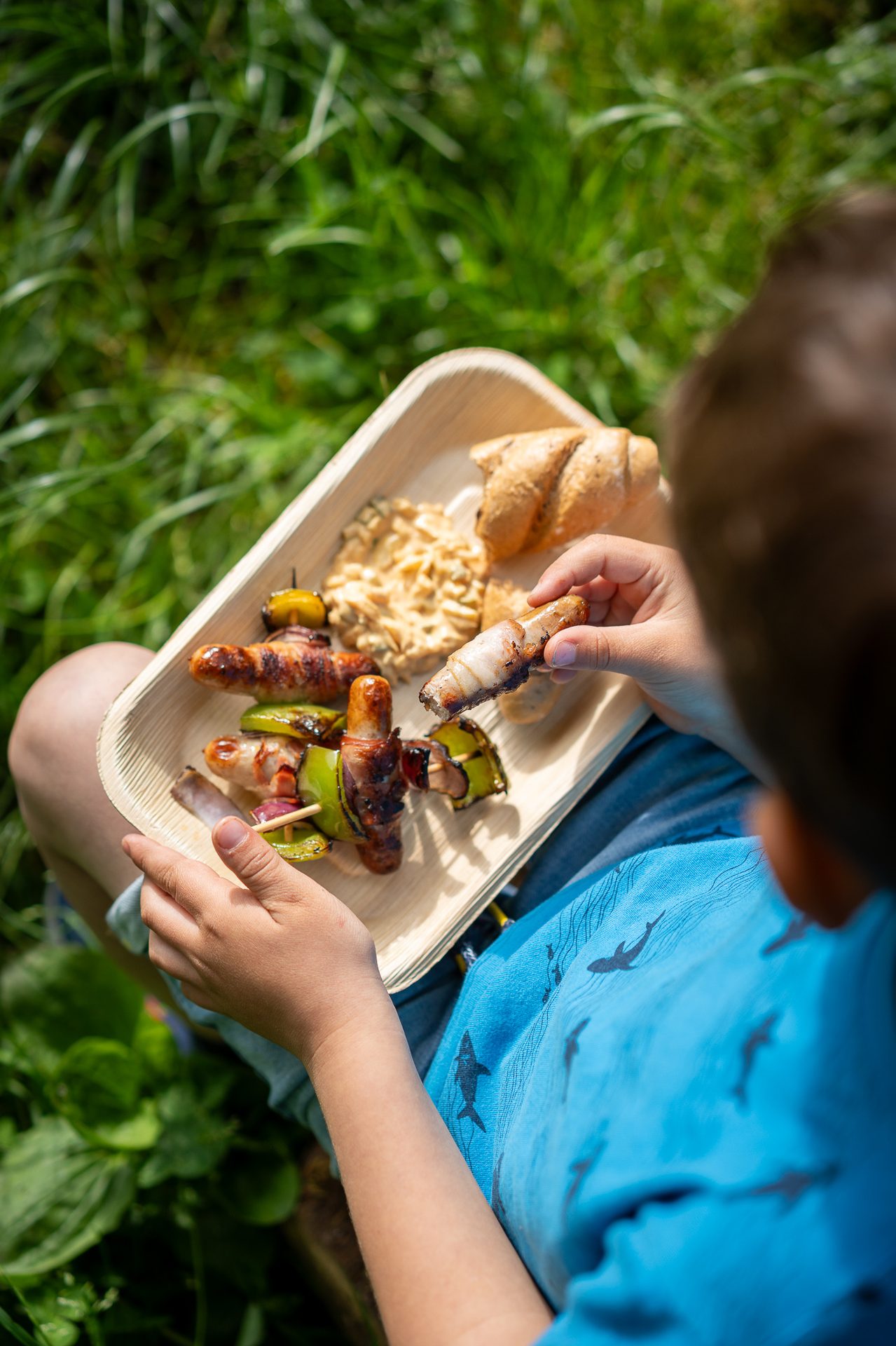 Child holding a plate with grilled skewers wrapped in bacon, bread, and dip, eating outdoors.