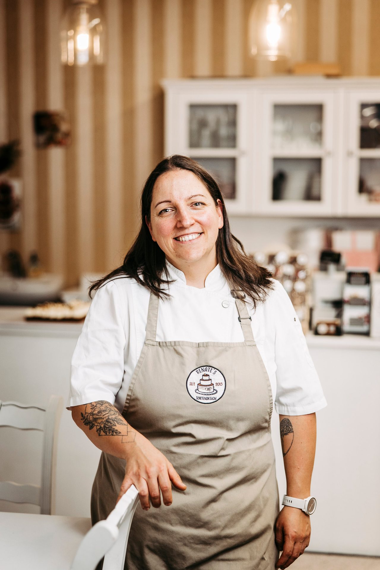 Smiling baker with tattoos in chef's uniform and "Renate's Tortendesign" apron.
