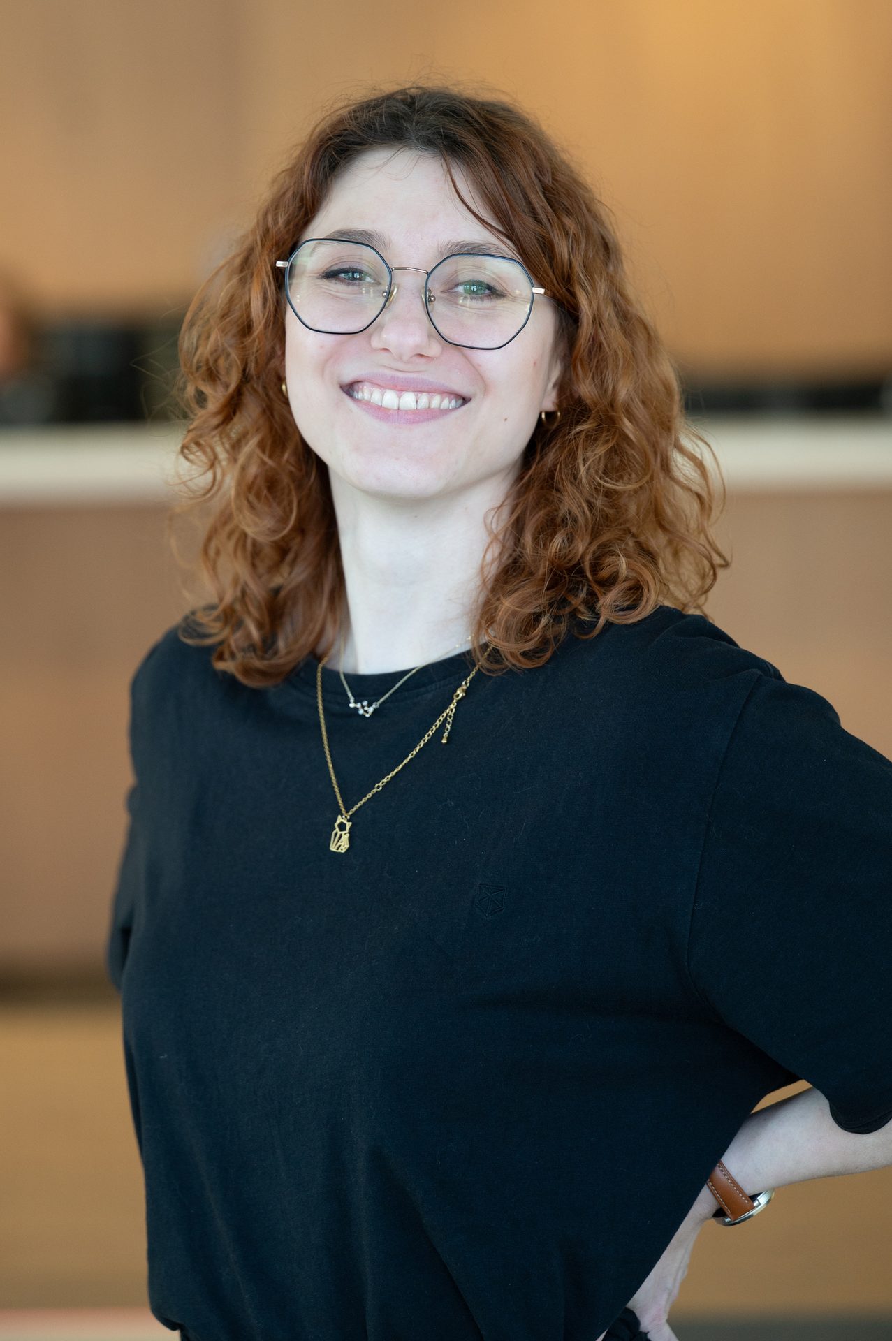 A smiling woman with red, curly hair and glasses, wearing a black t-shirt and necklaces.