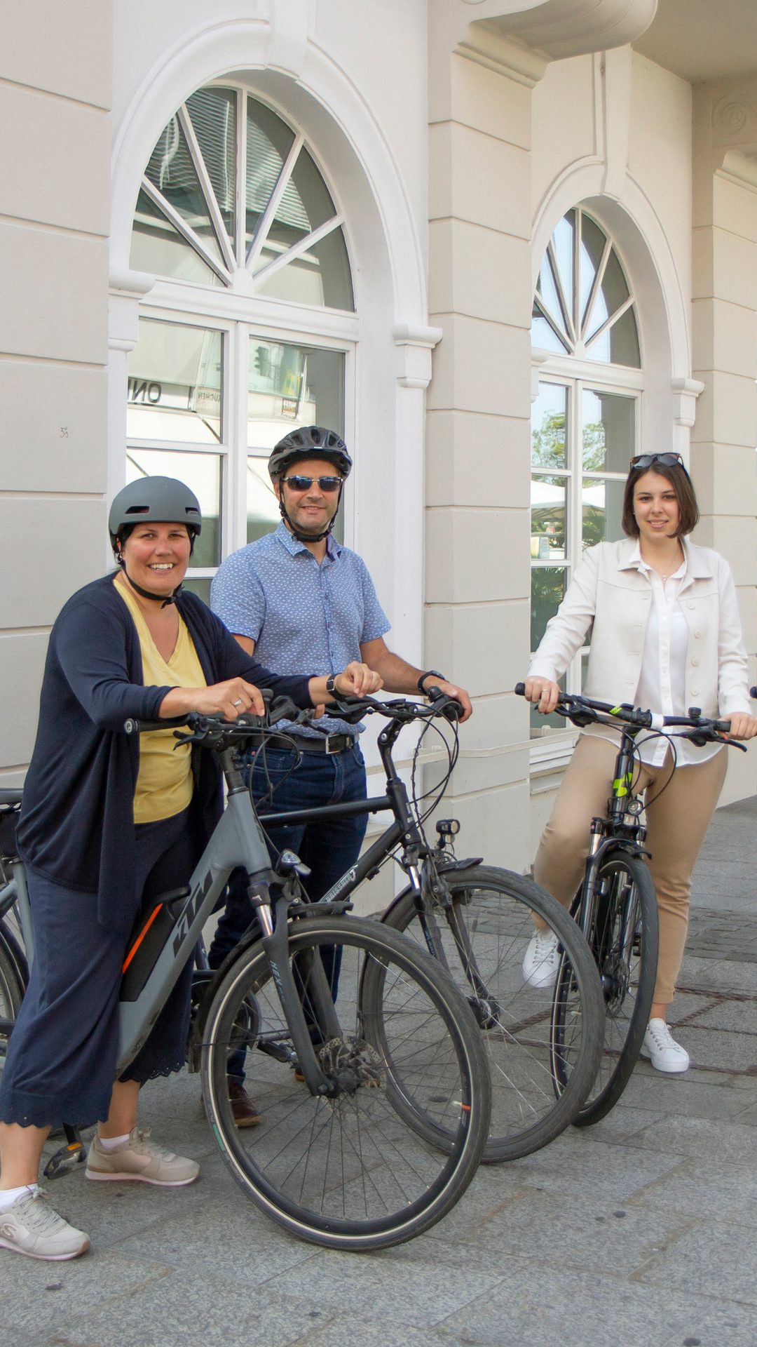 Two men and one woman stand with bicycles outside a building.
