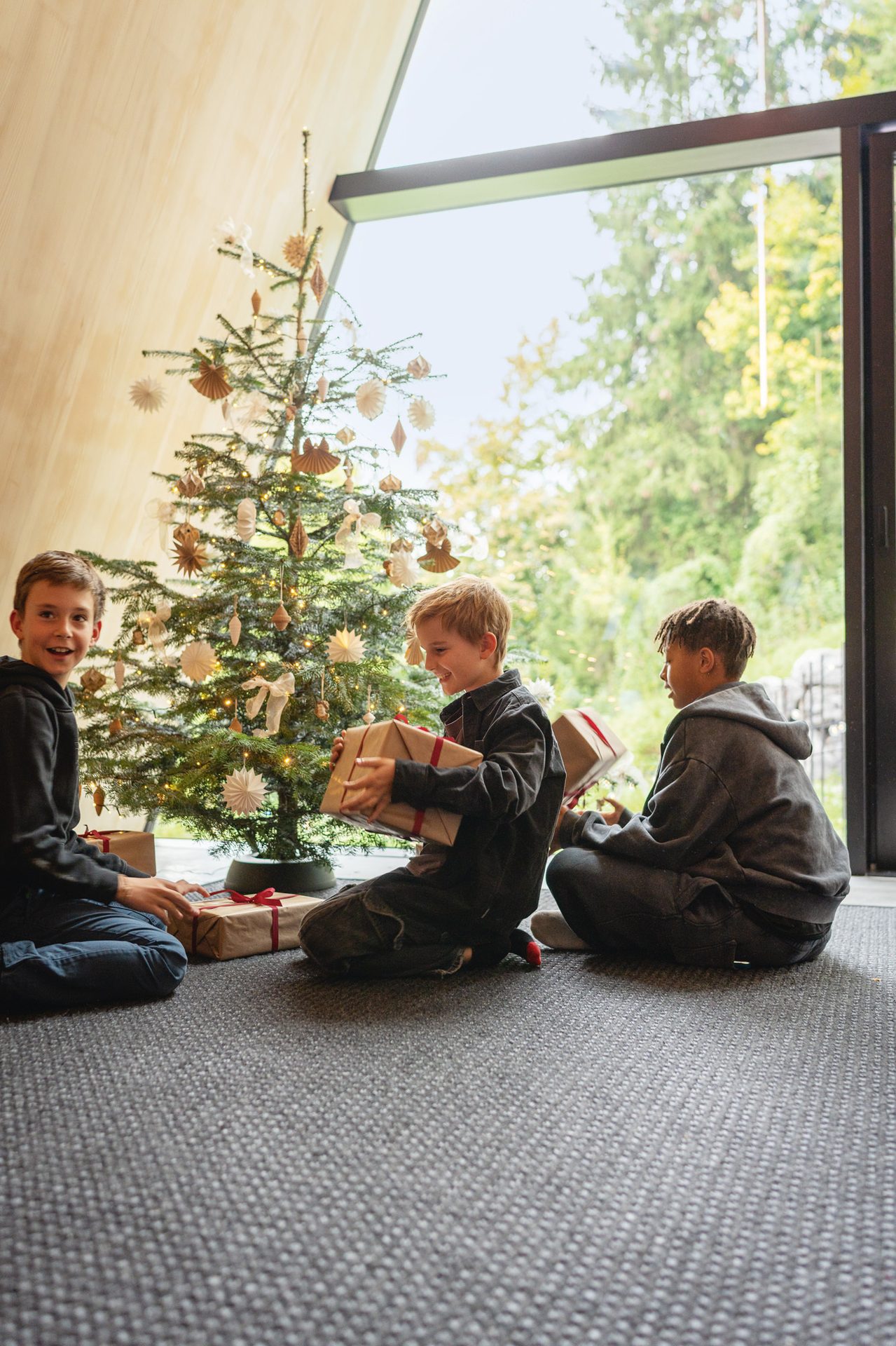 Three boys with gifts around a Christmas tree, sitting on a carpet near a window.