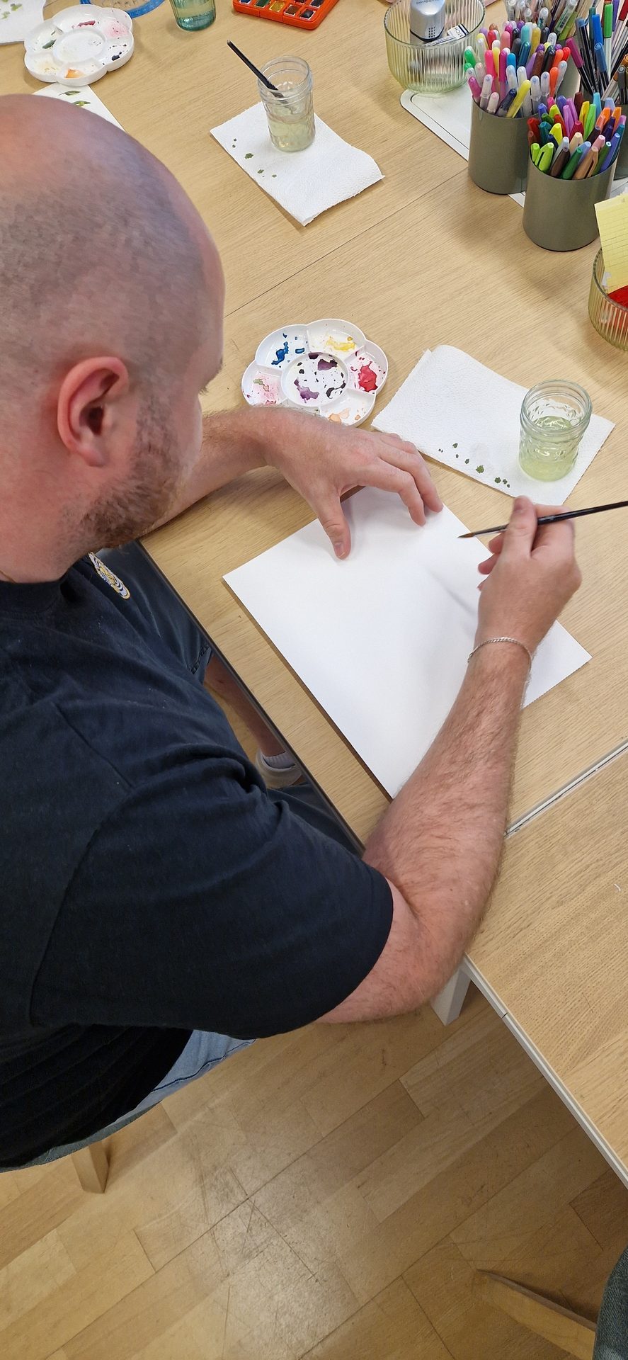 Person painting with watercolors on white paper at wooden table, overhead view.