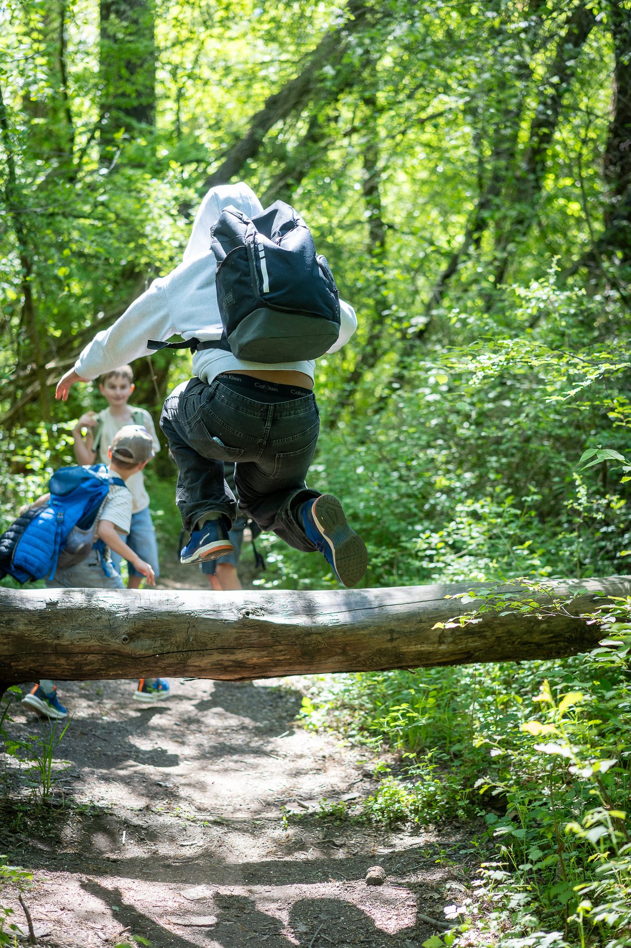 A child with a backpack leaps over a log on a forest trail, with other kids behind.