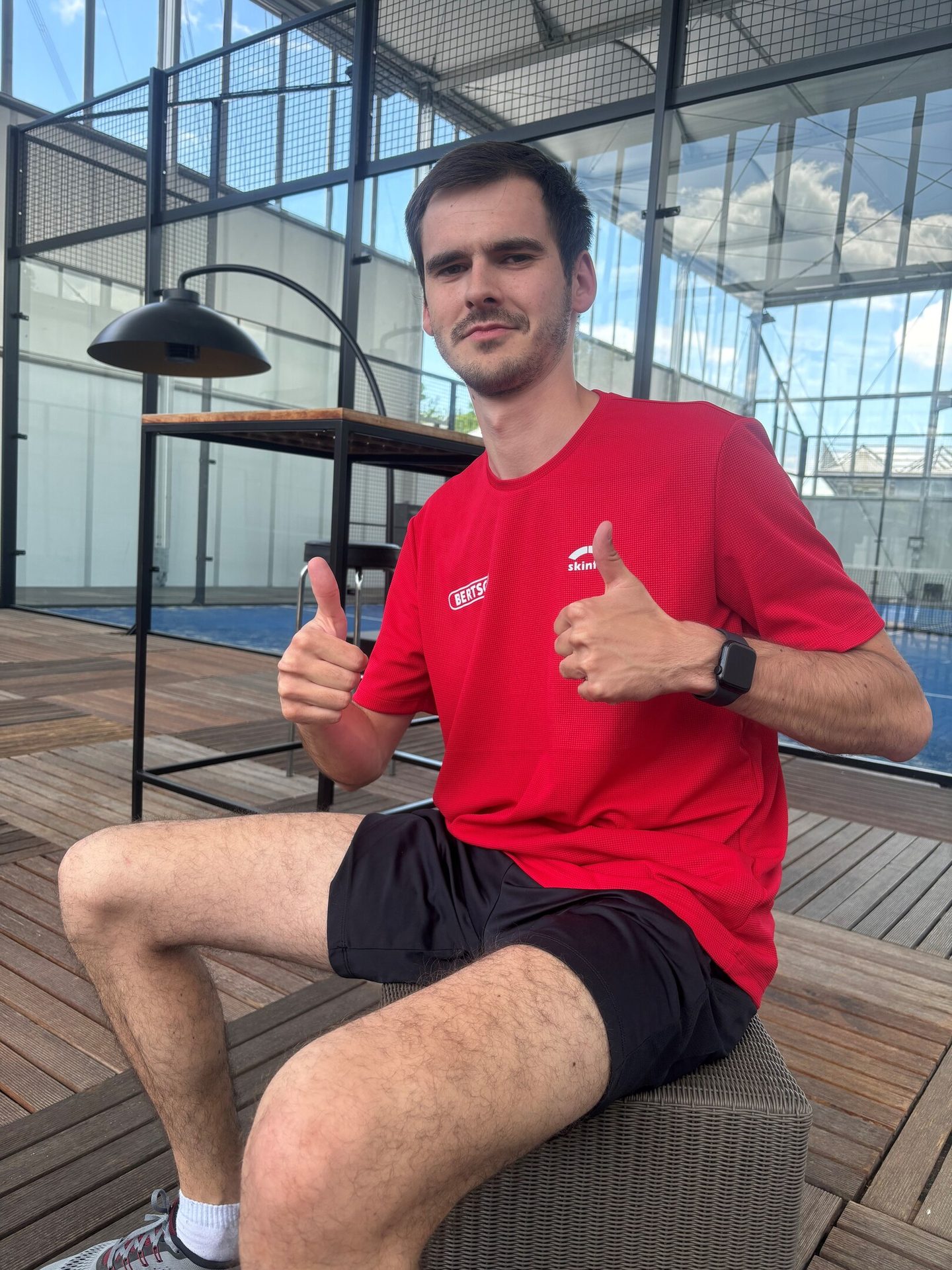 Man in red shirt and shorts sitting outdoors, giving two thumbs up.