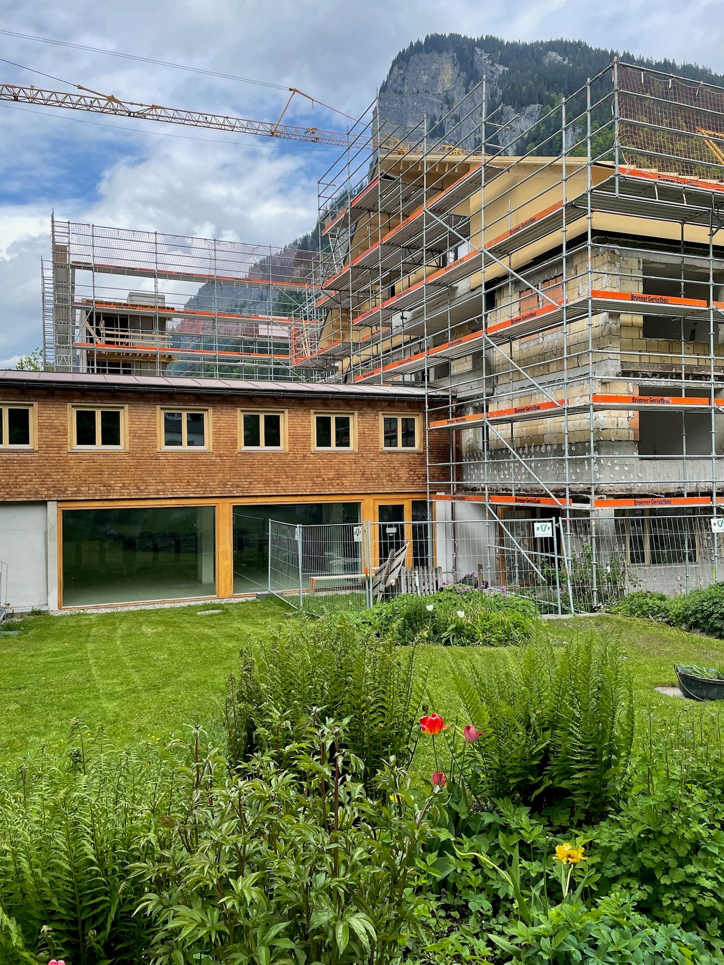 Building under construction with scaffolding and crane, mountain backdrop, and a garden with flowers.