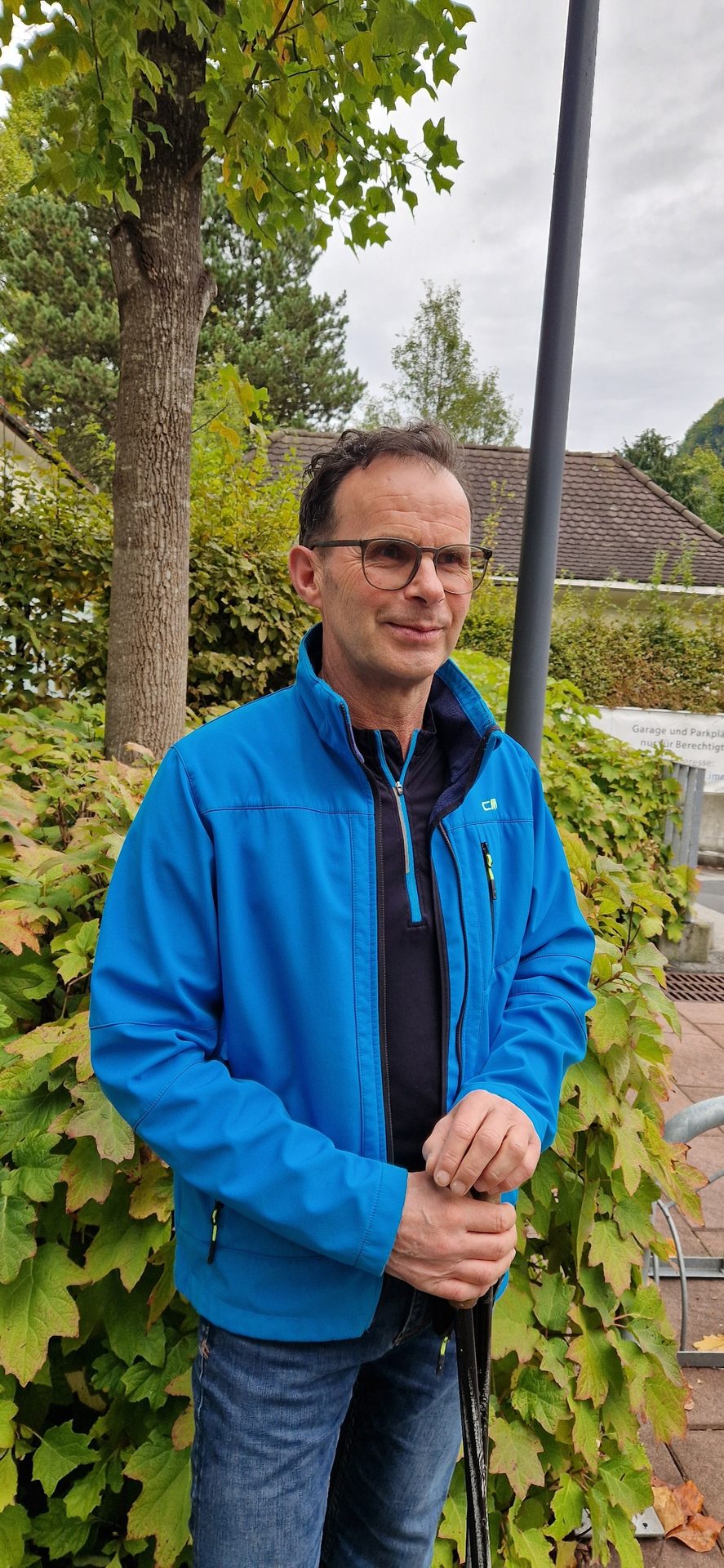 Smiling man in blue jacket and glasses, holding a stick outdoors.