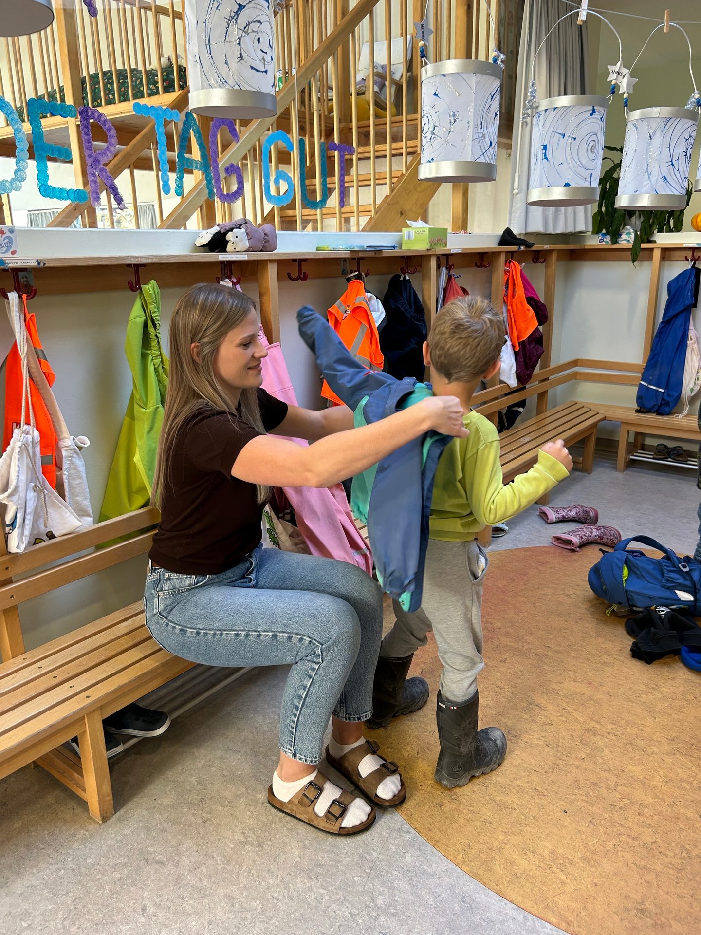 A woman helps a young child put on a jacket in a brightly lit room with cubbies and benches.