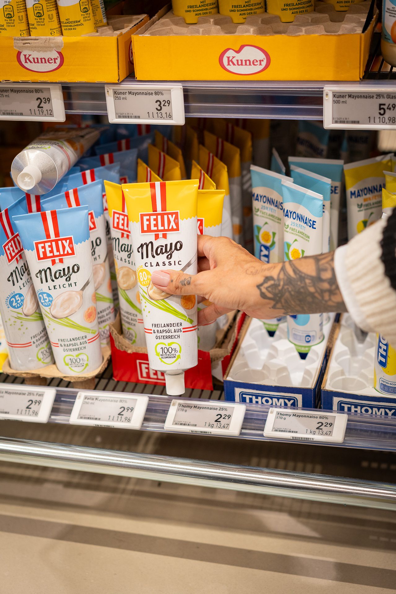 A person's hand reaches for a tube of Felix Mayo Classic mayonnaise in a supermarket aisle. Other mayo brands are visible.