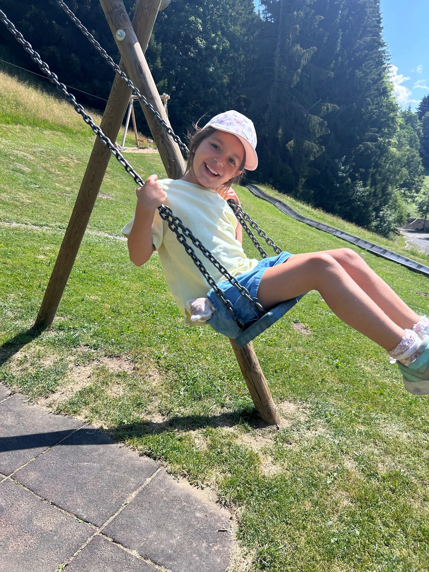 A happy young girl in a white cap and yellow shirt smiling while swinging outdoors on a sunny day.