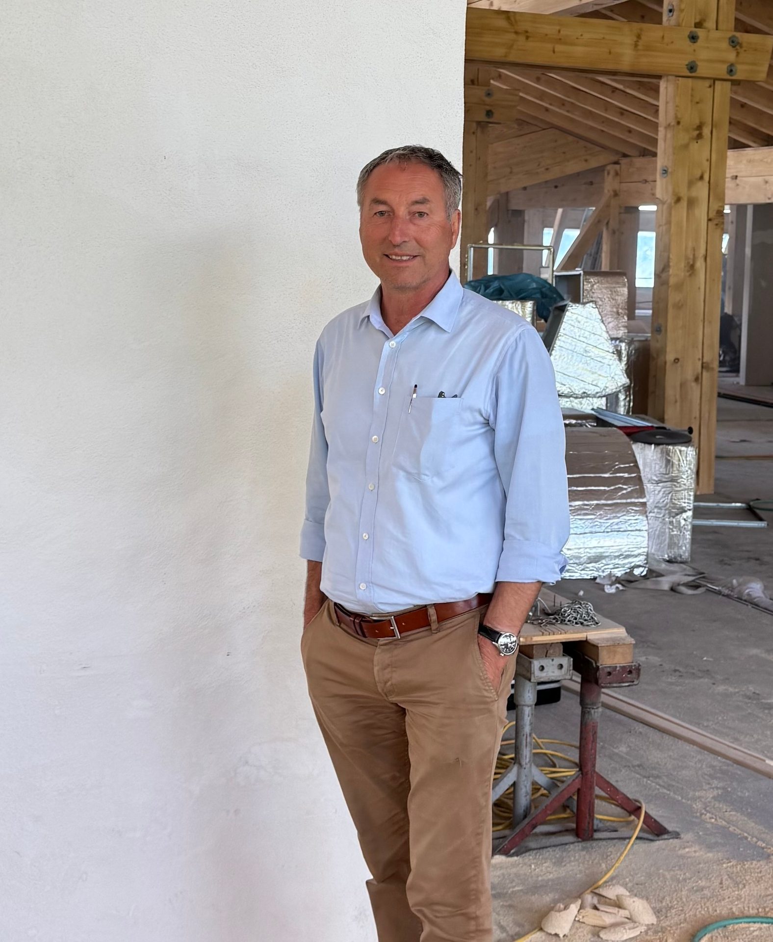 A smiling man in a blue shirt and brown pants stands with hands in pockets at a construction site.