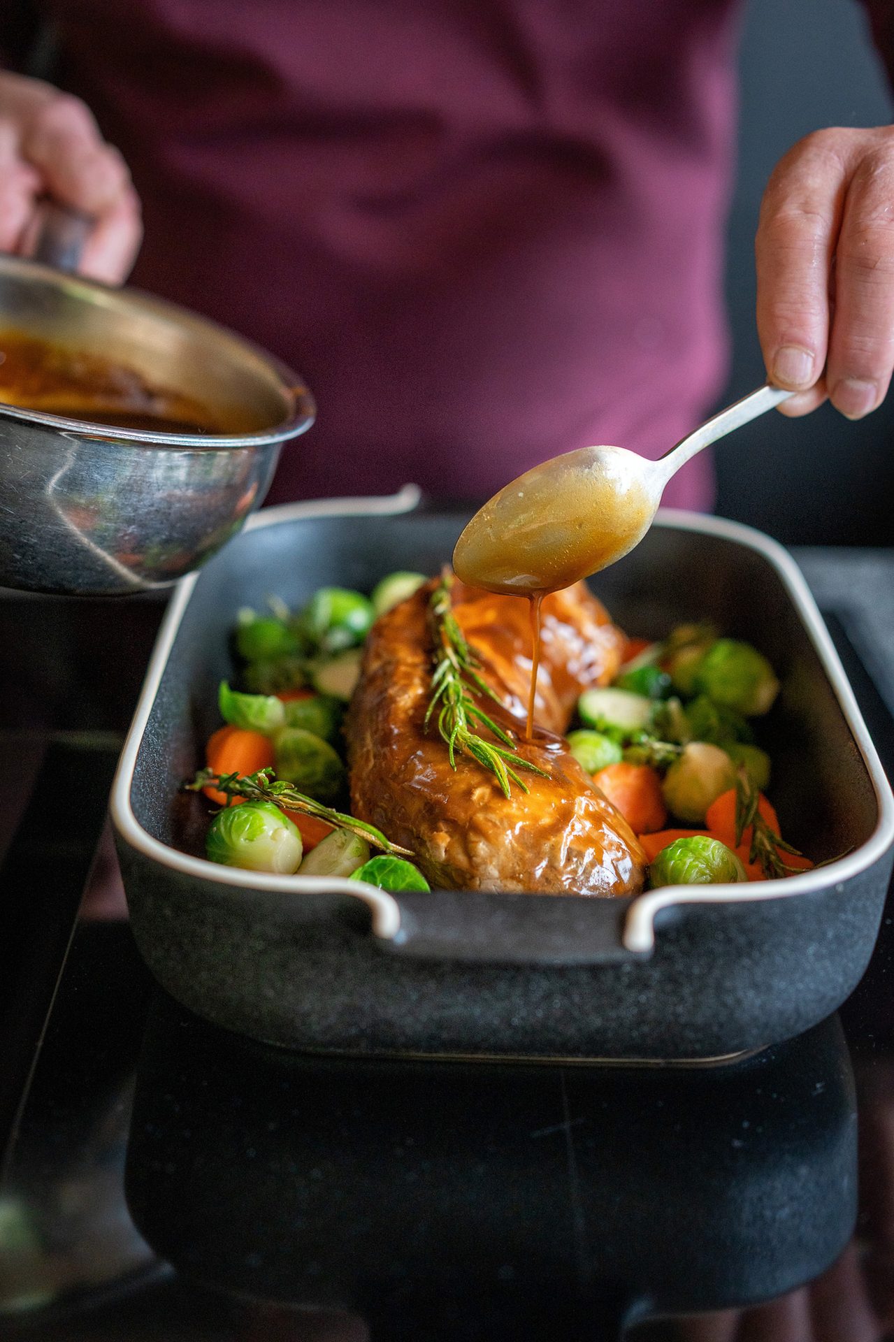 A hand spoons gravy over a glazed roast with brussels sprouts and carrots in a dark baking dish.