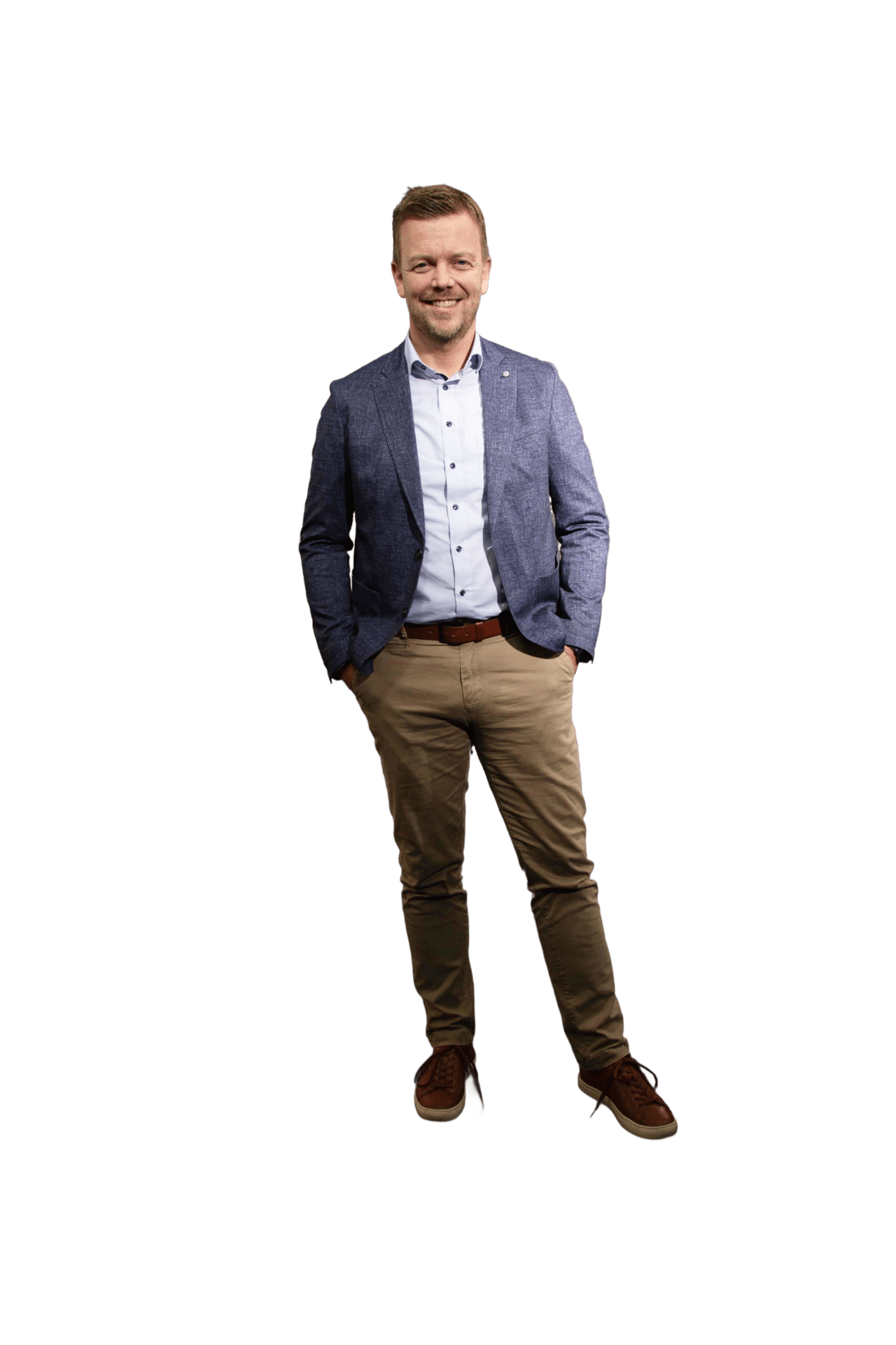 A smiling man in a blue blazer, light shirt, khaki pants, and brown shoes, with hands in pockets.
