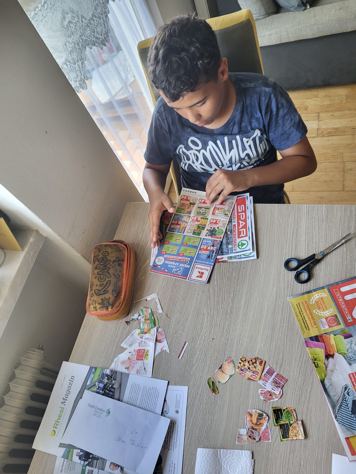A boy cuts pictures from a flyer at a table with scissors and other craft materials.