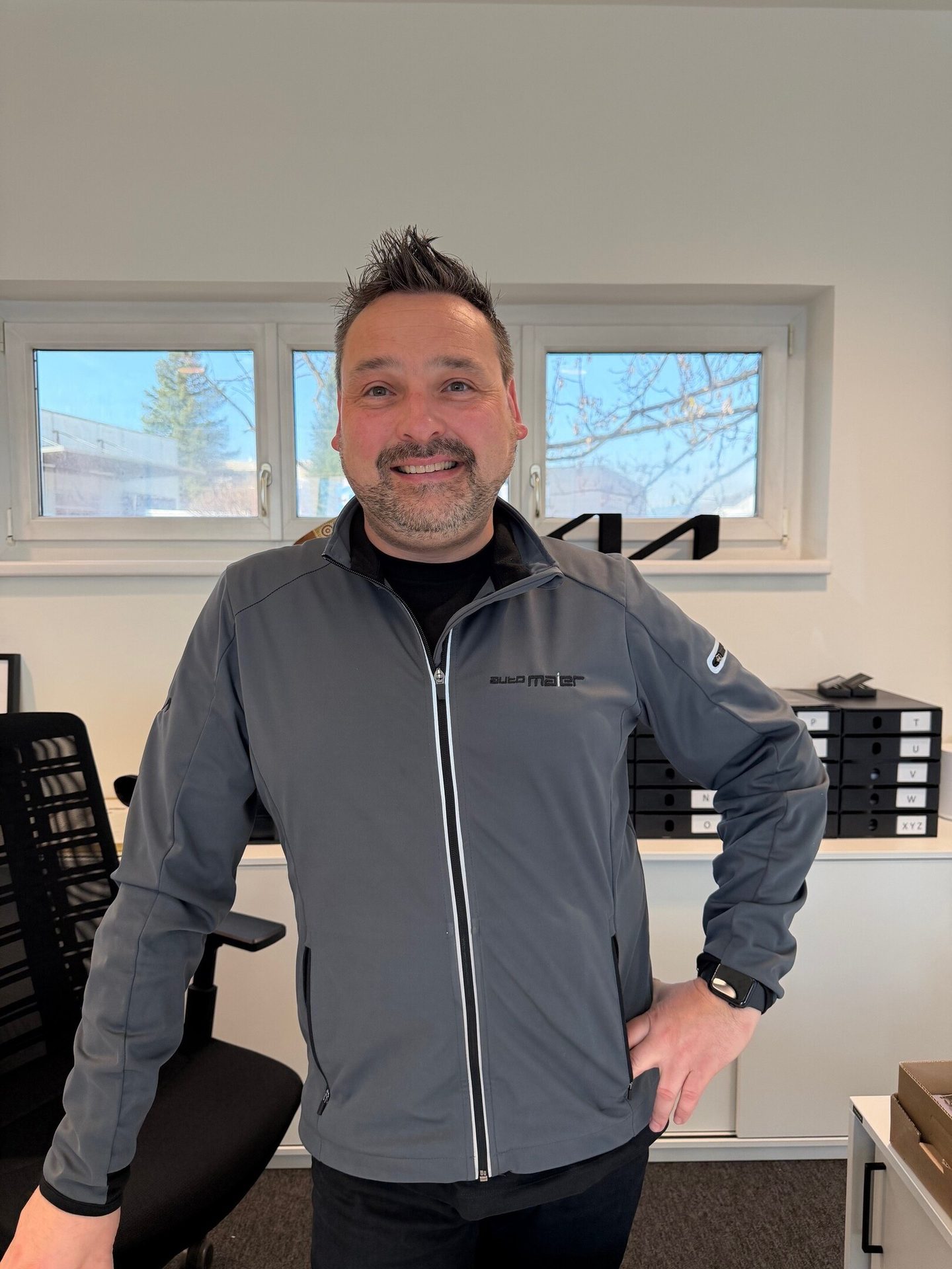 A smiling man in a gray "auto Maier" jacket stands confidently in an office setting with windows.