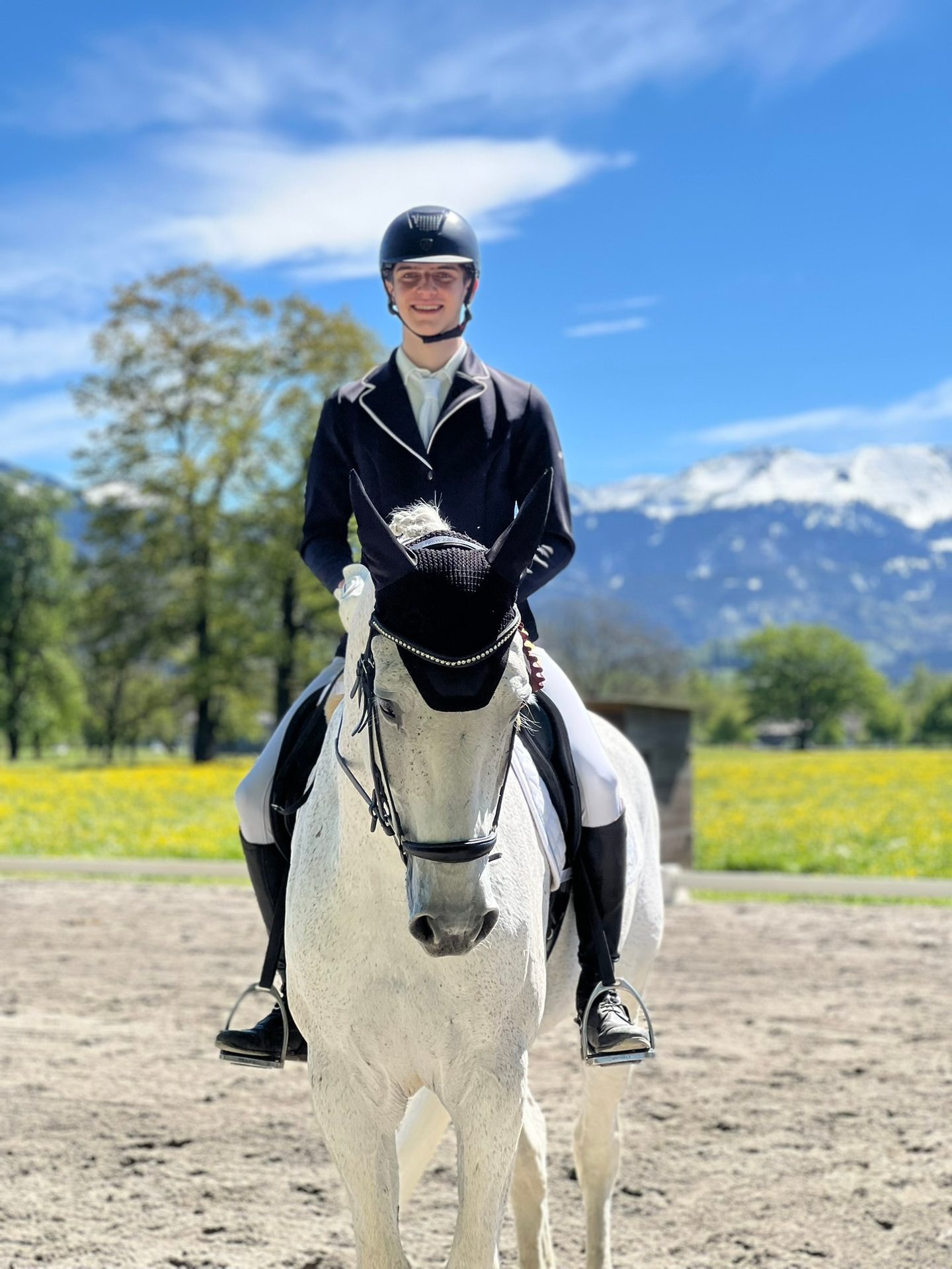 Smiling rider on a white horse, dressed in riding attire, with snowy mountains and green fields.