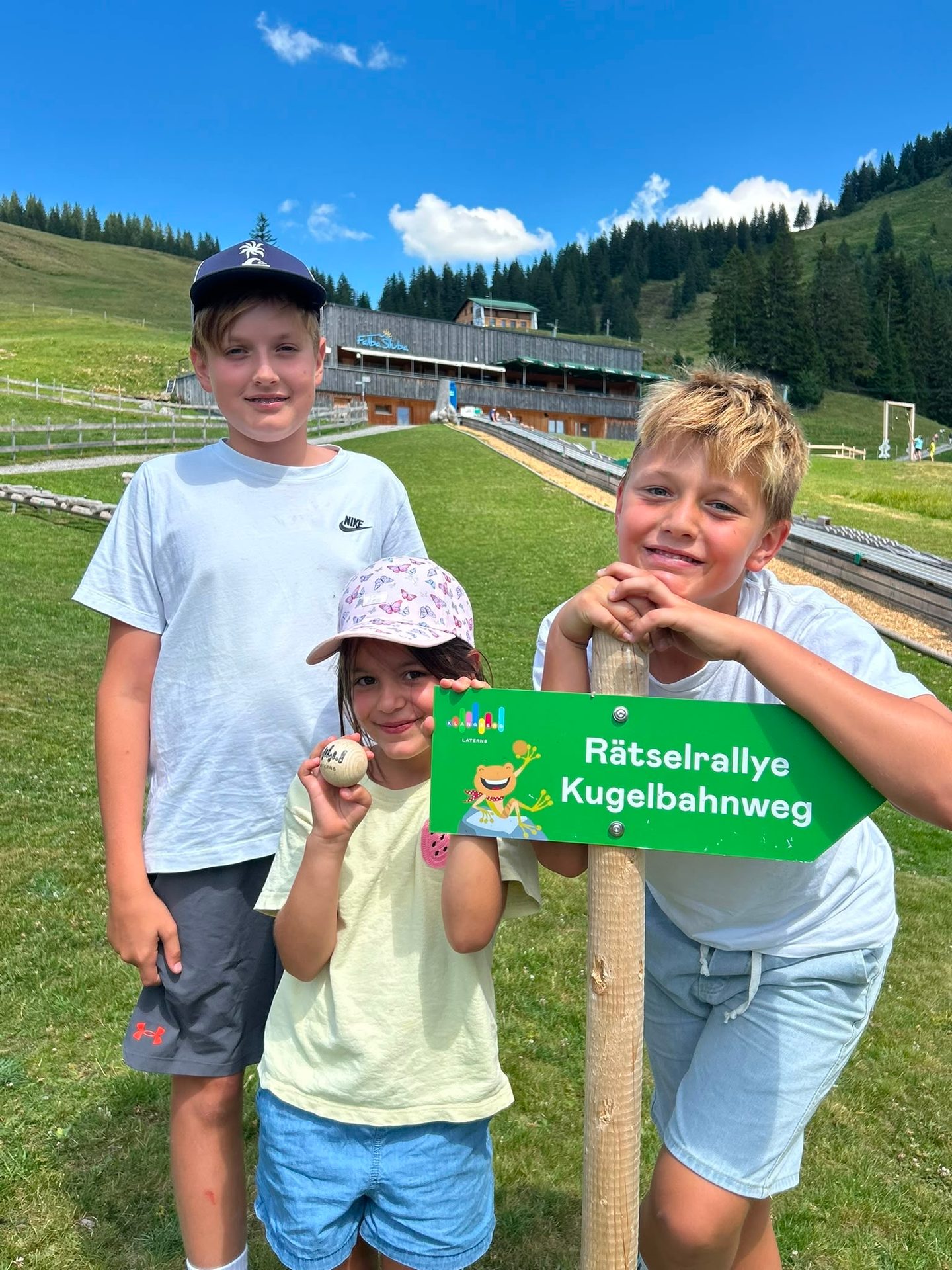 Three children pose with a sign for the "Rätselrallye Kugelbahnweg" on a sunny mountain day.