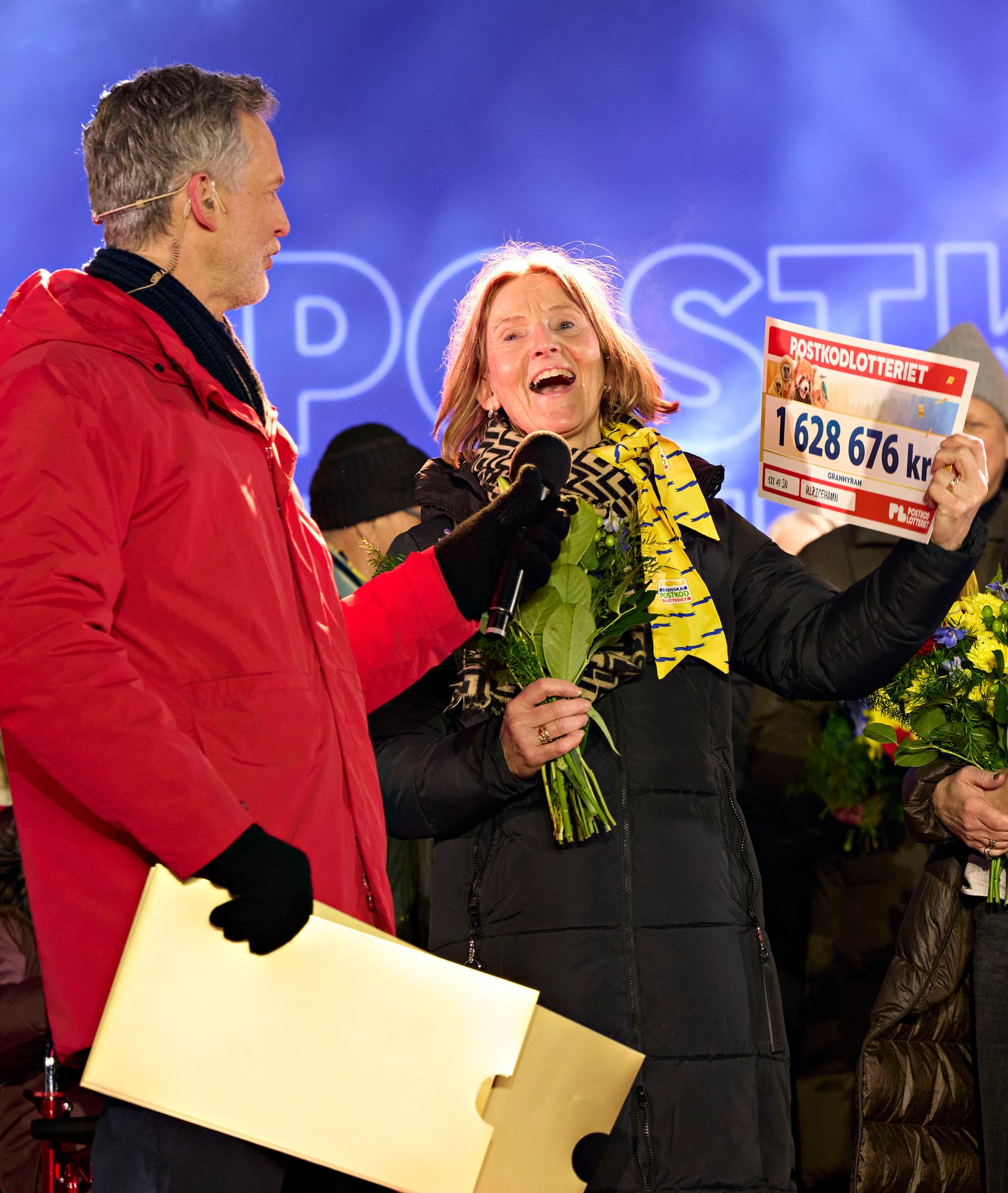 Excited woman holds a 1.6M kr lottery check and flowers on stage with a man holding a mic.