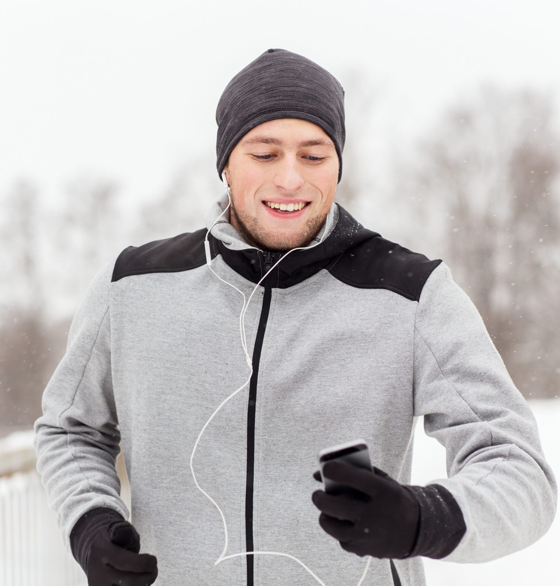 A happy man in winter running gear with earphones checks his phone while jogging in the snow.
