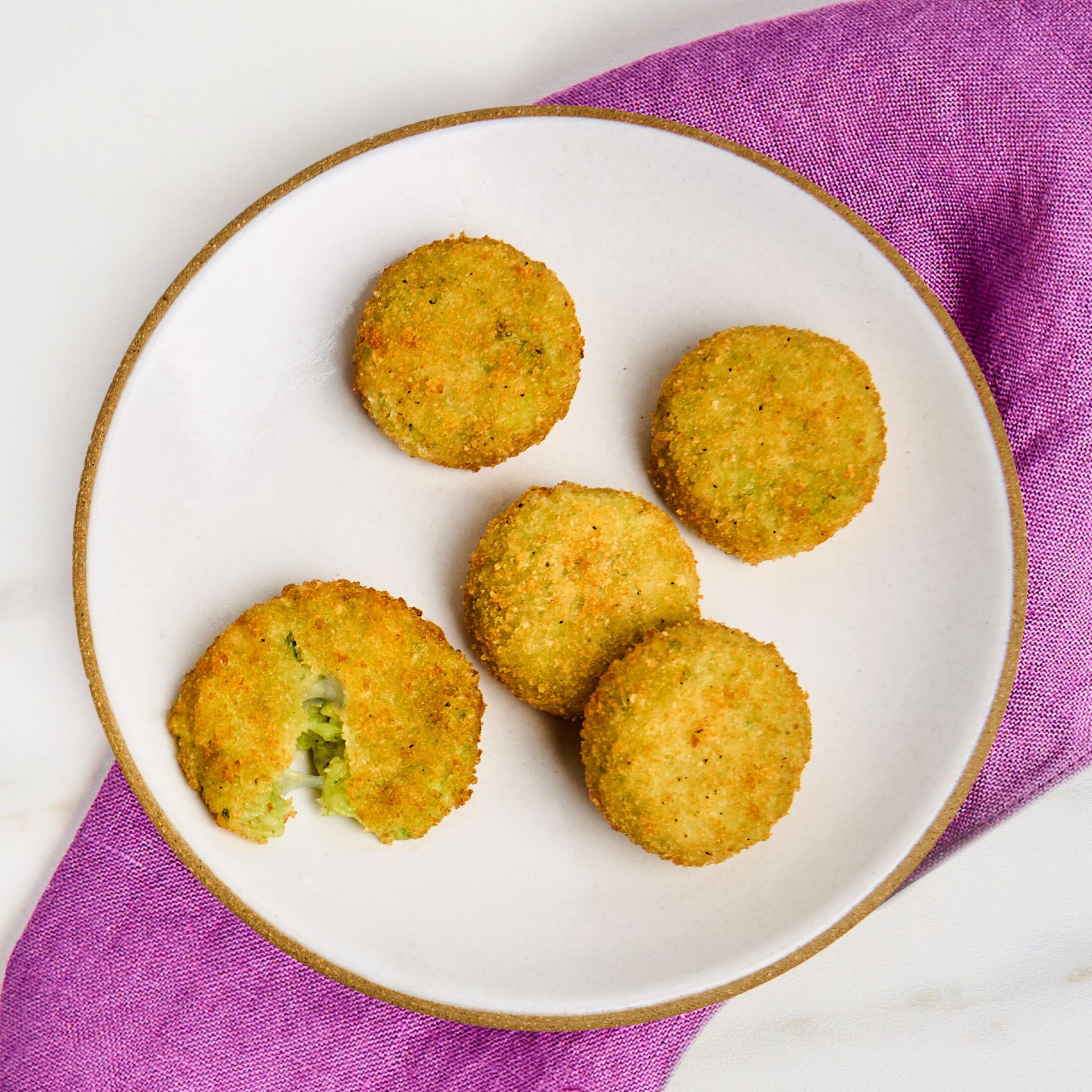 Five breaded green broccoli & cheese bites on a white plate; one broken shows gooey interior. Purple napkin.