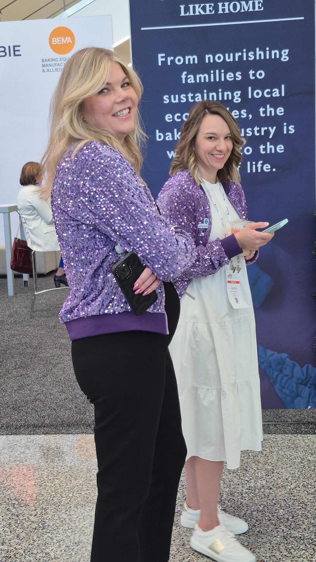 Two smiling women in purple sequin jackets at a conference, holding phones.