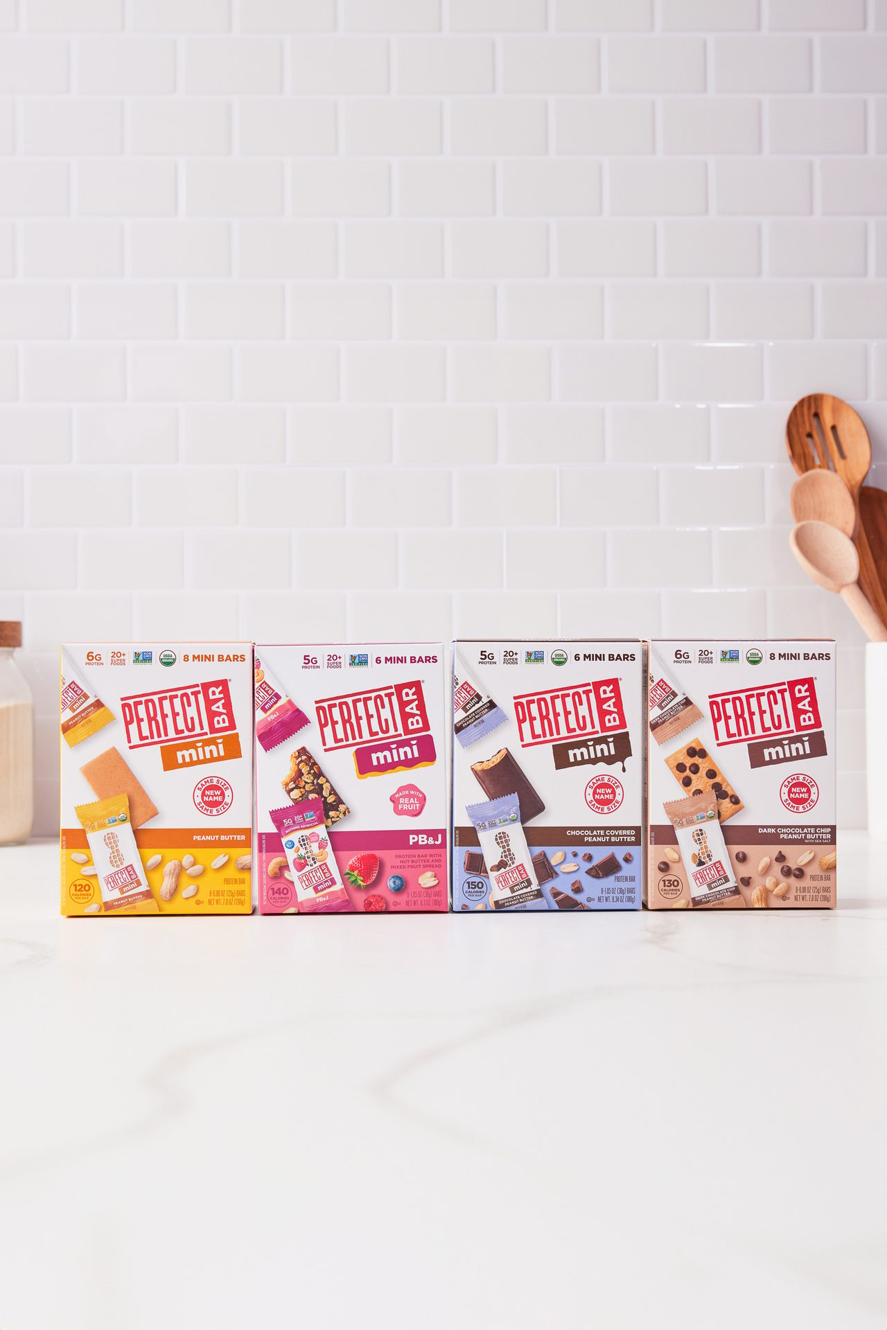 Four boxes of Perfect Bar Mini protein bars in various flavors lined up on a white kitchen counter.