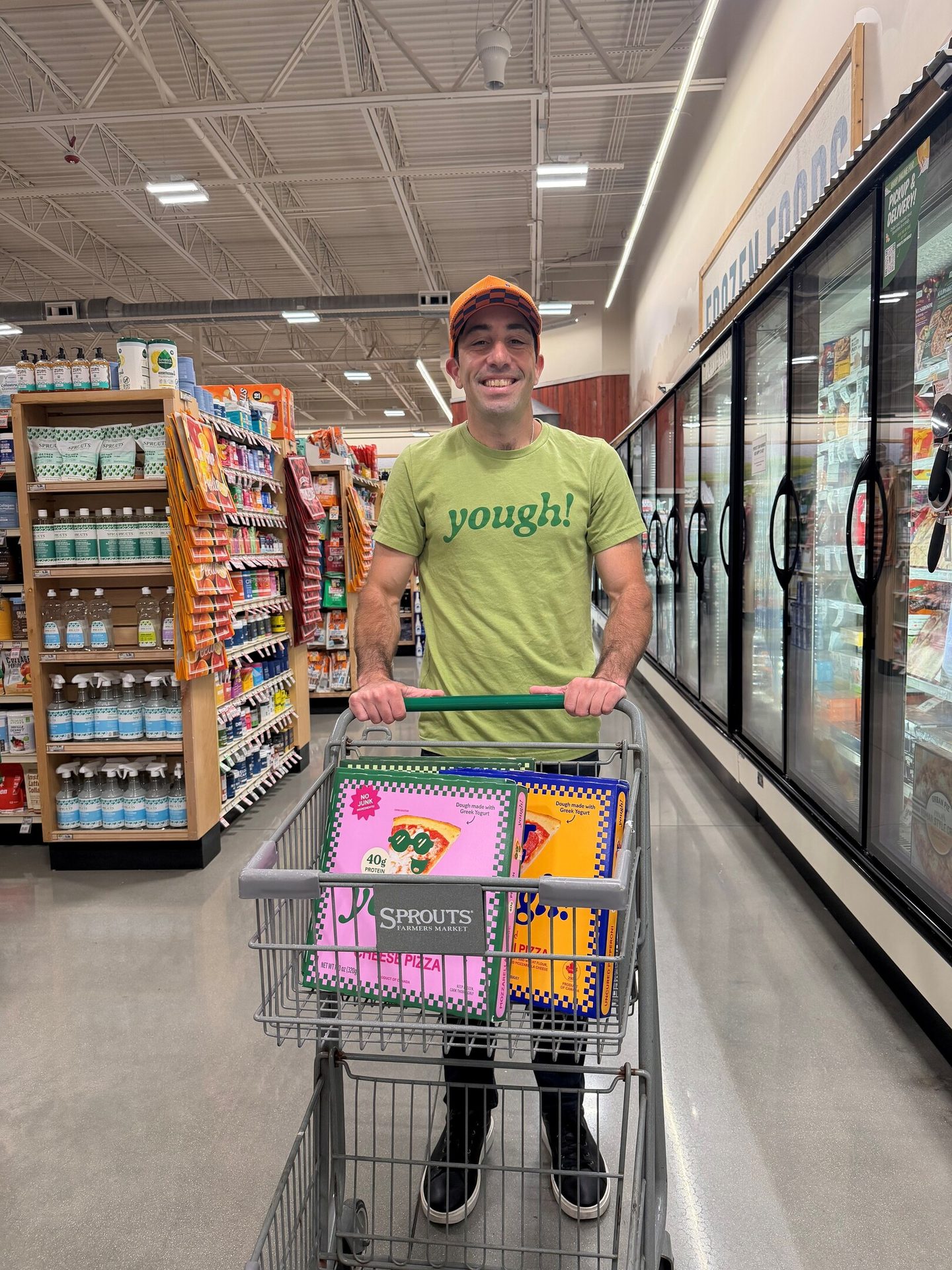 A smiling man pushing a grocery cart with pizza boxes in a supermarket aisle.
