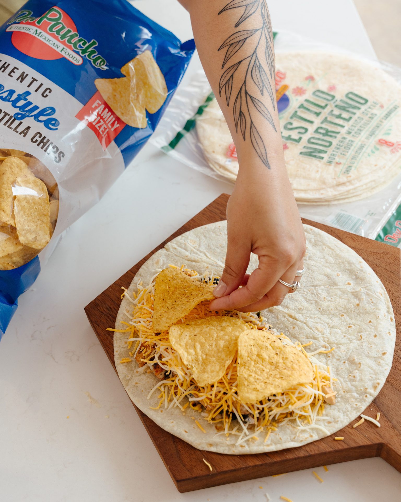 Person adding tortilla chips to a flour tortilla filled with cheese and other ingredients on a wooden board.