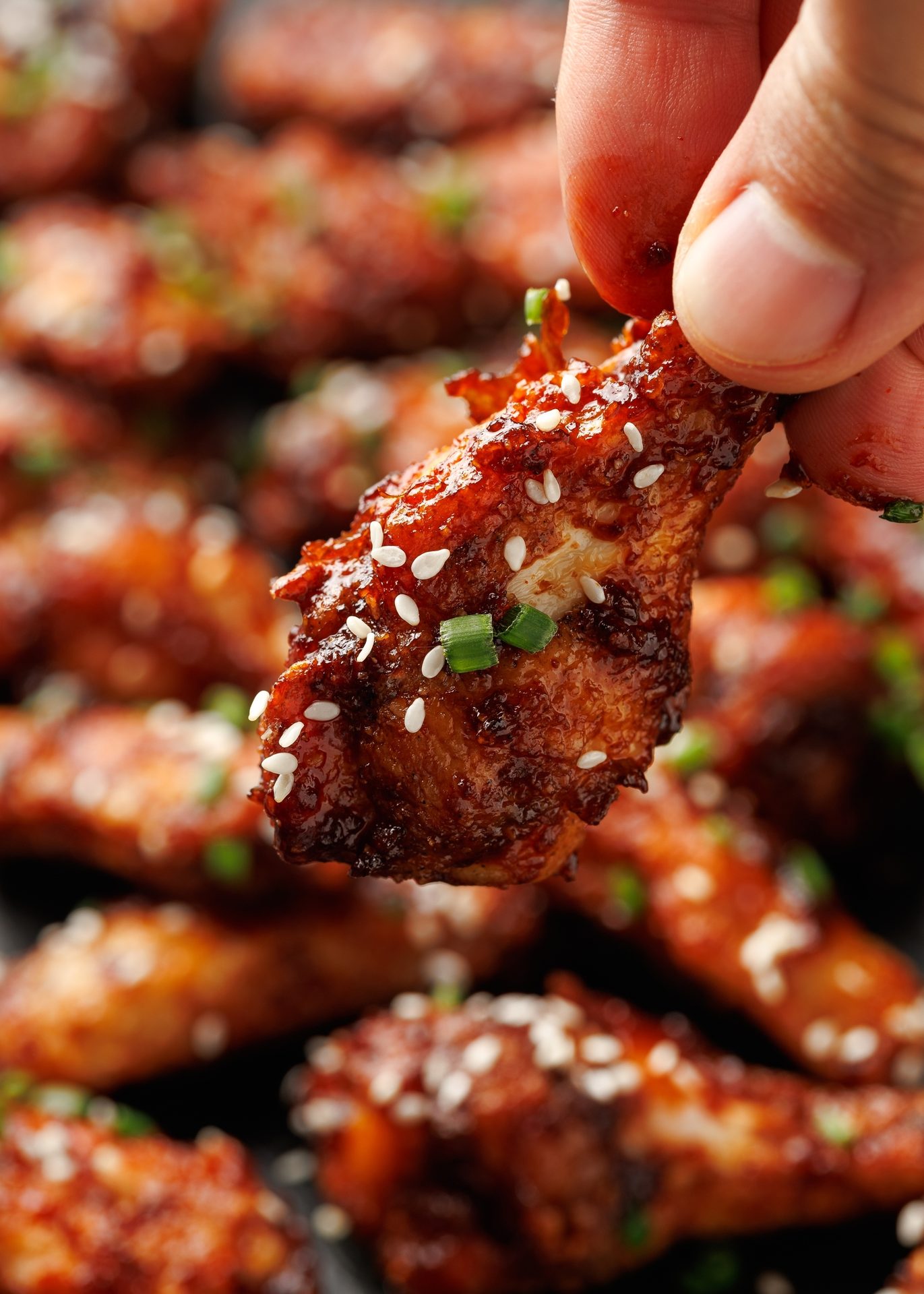 A hand holds a sesame-crusted, glazed chicken wing, with more wings blurred in the background.