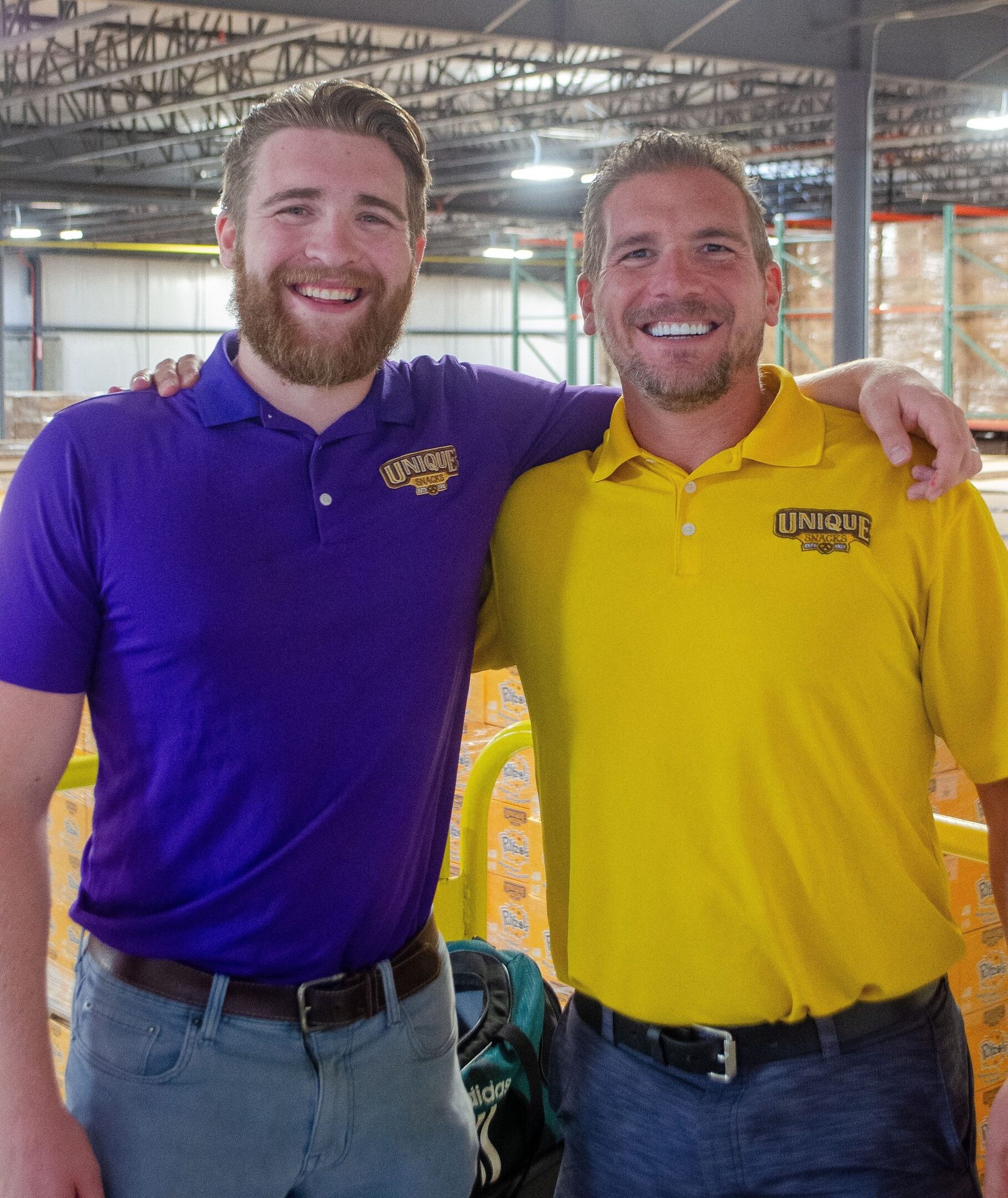 Two smiling men in "Unique Snacks" purple and yellow polos, embracing in a warehouse.