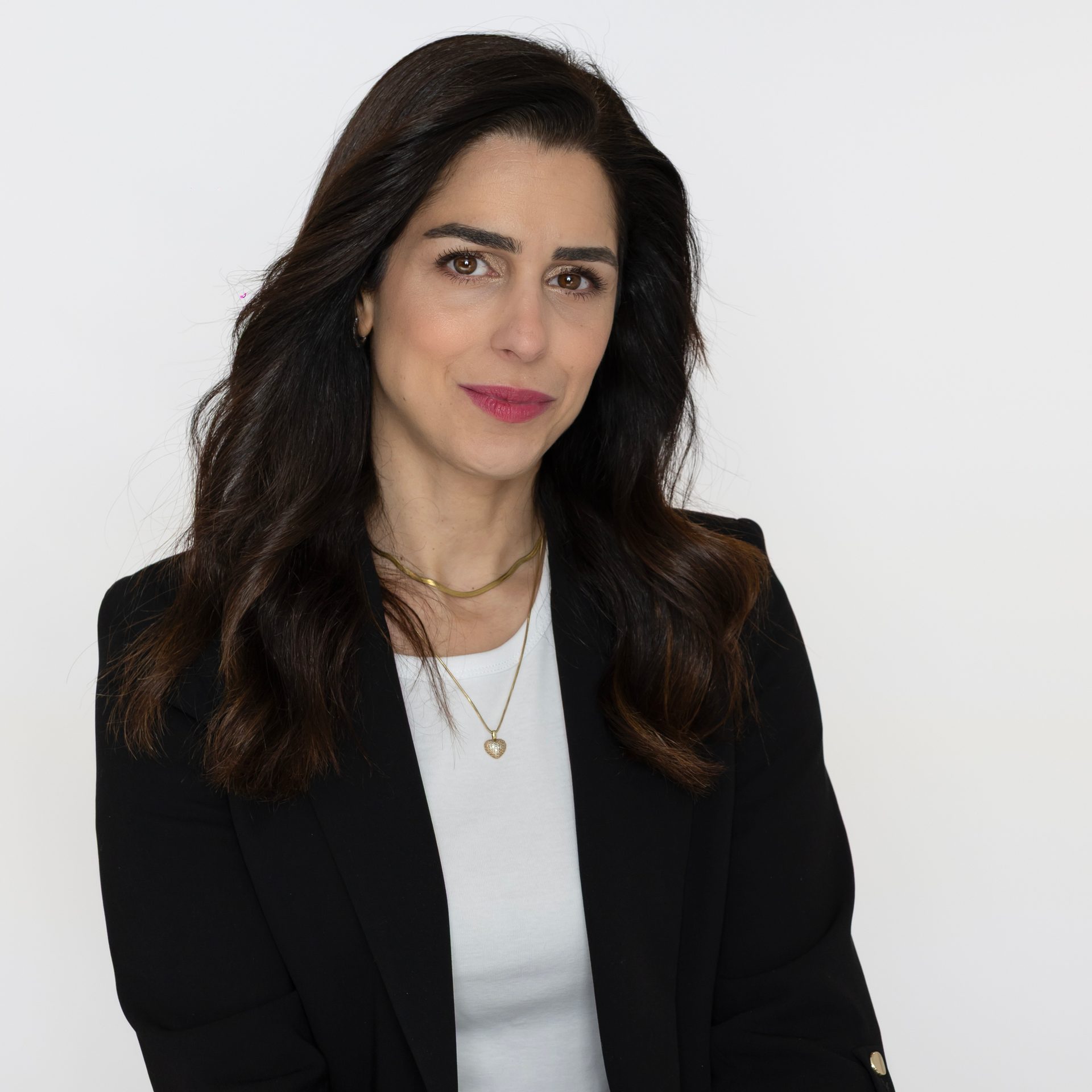 Headshot of a woman with long dark hair, black blazer, white top, and gold necklaces.