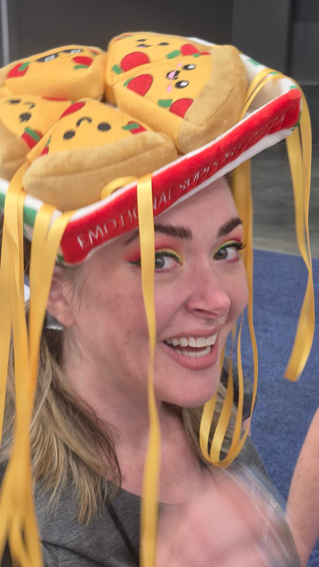 Smiling person in a pizza hat with yellow ribbons, vibrant eye makeup.