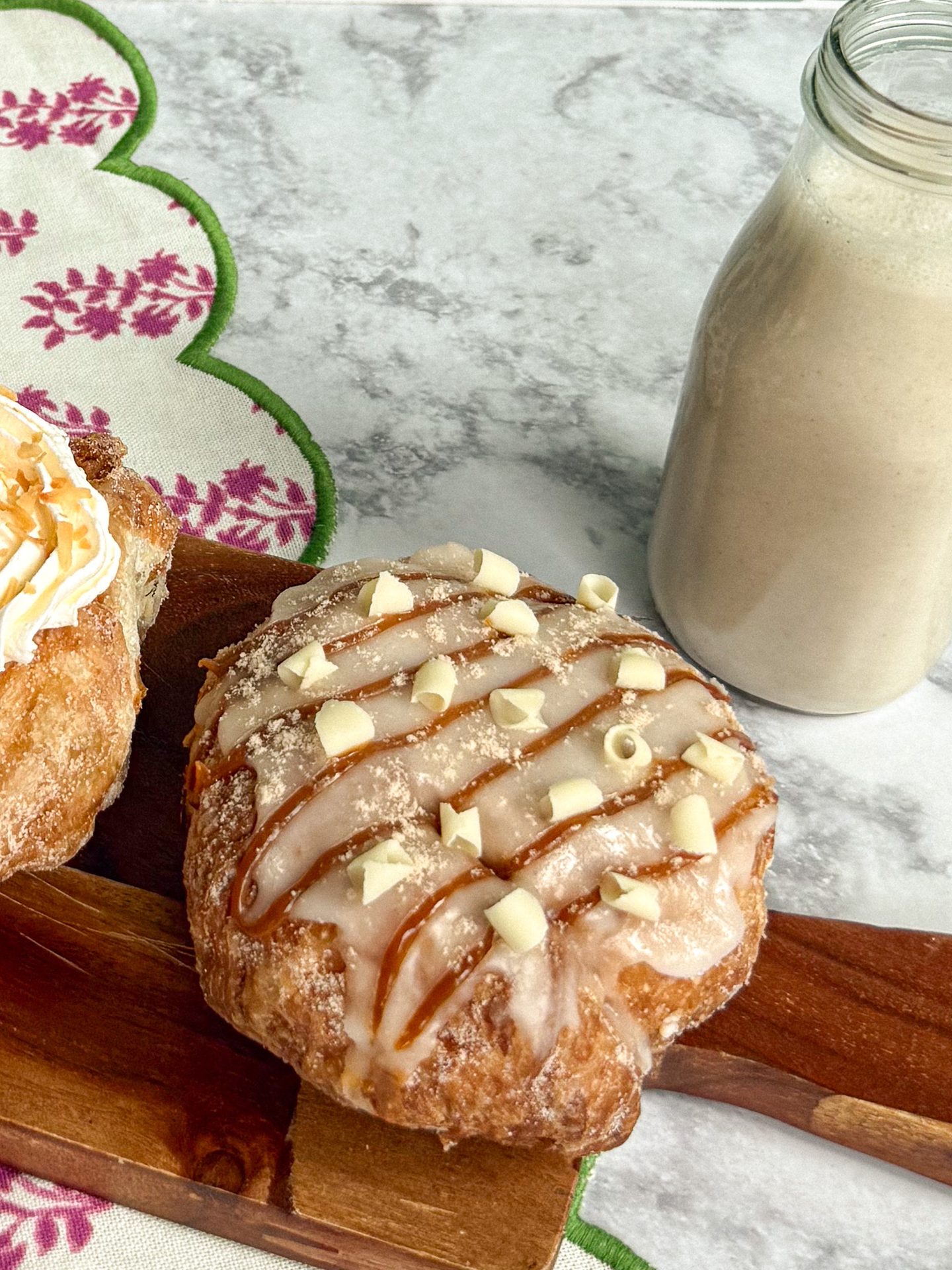 A glazed donut with caramel drizzle and white chocolate shavings next to a bottle of milk on a wooden board.