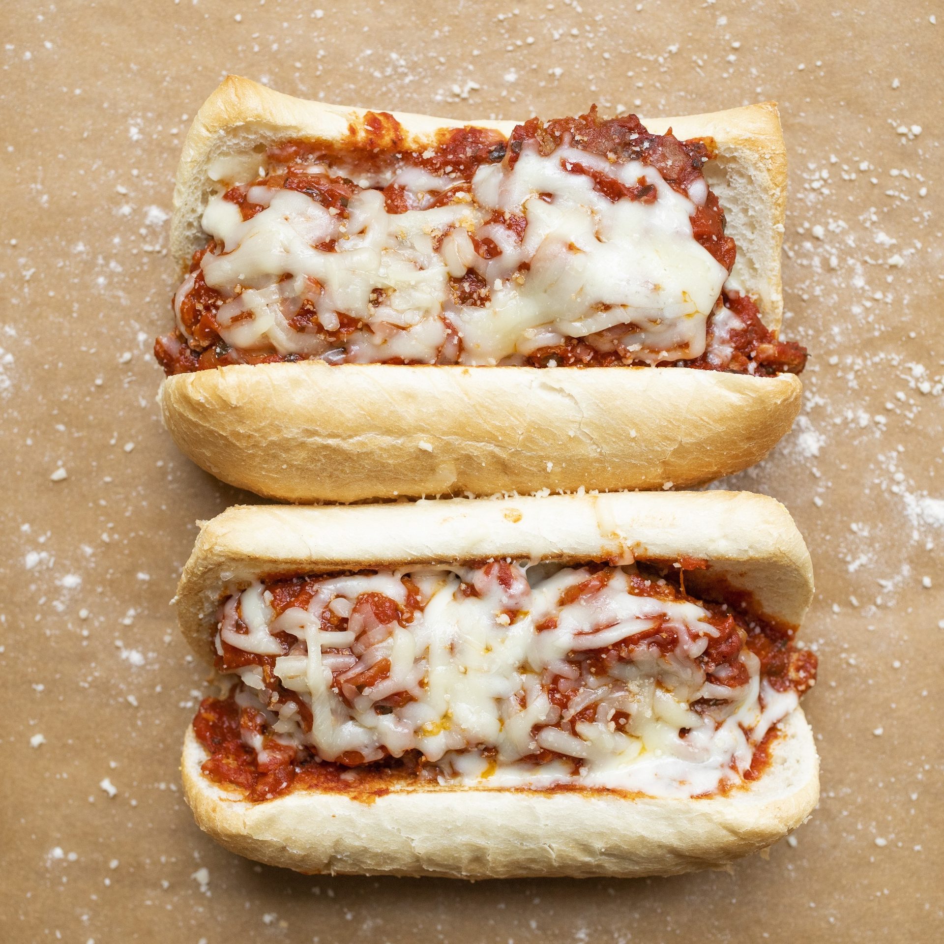 Two cheesy meatball subs on a brown surface with white flecks.