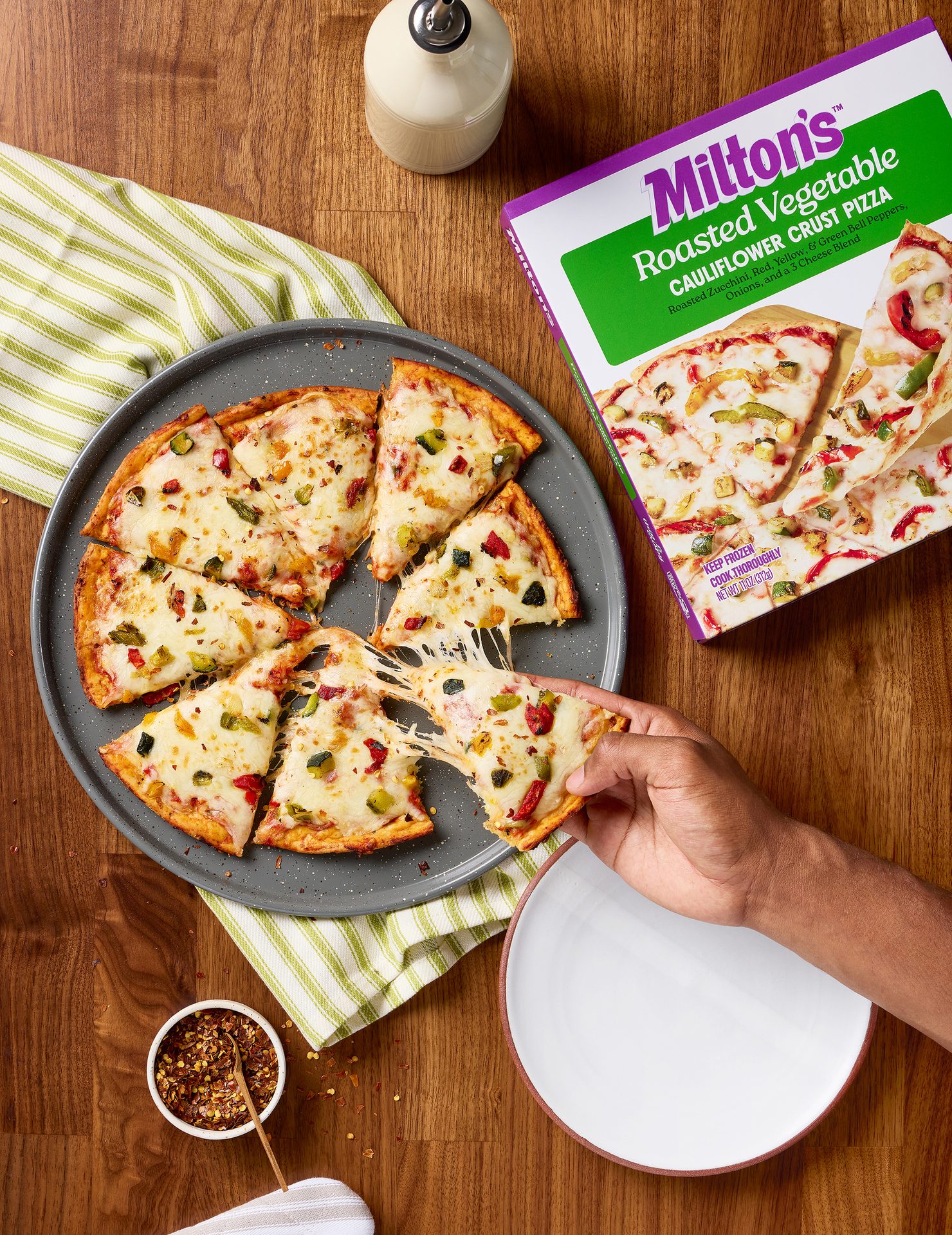 Milton’s cauliflower crust roasted vegetable pizza on a pizza pan, with someone pulling a slice