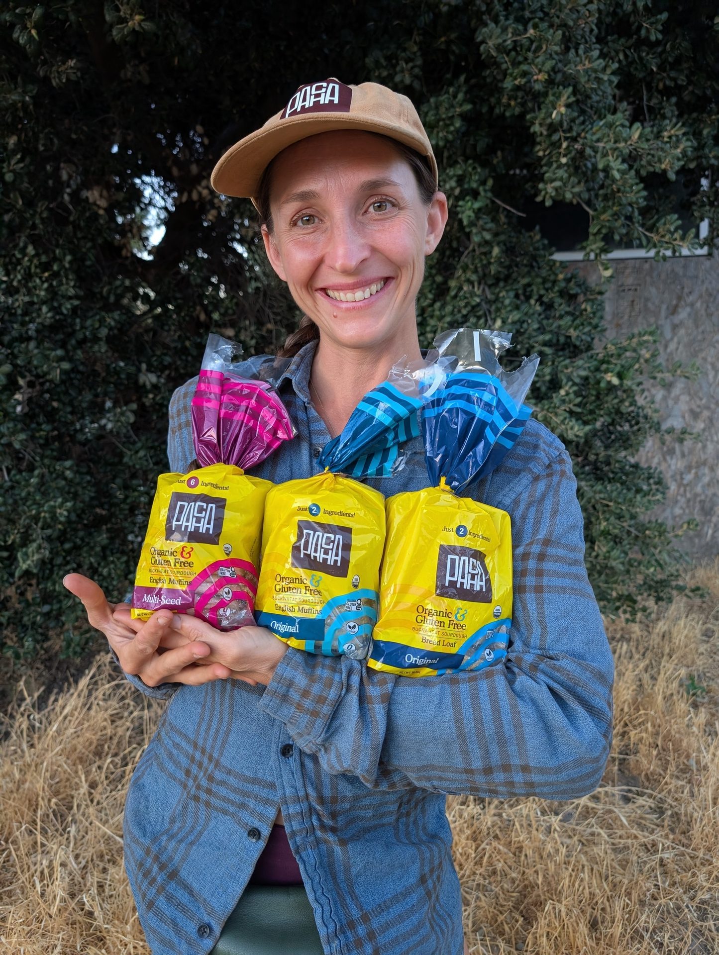 A smiling woman wearing a hat holds three yellow "PASA" brand organic & gluten-free bread product bags.