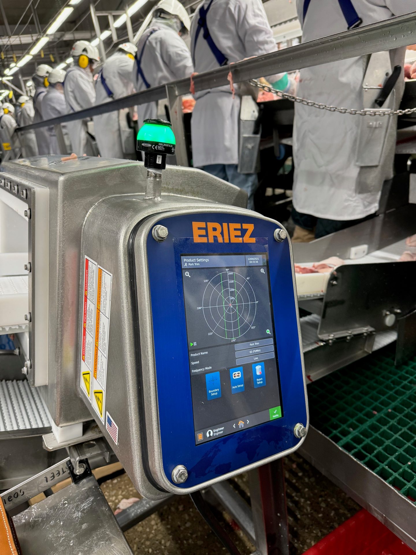 ERIEZ metal detector screen in a meat processing plant, with workers and machinery in the background.
