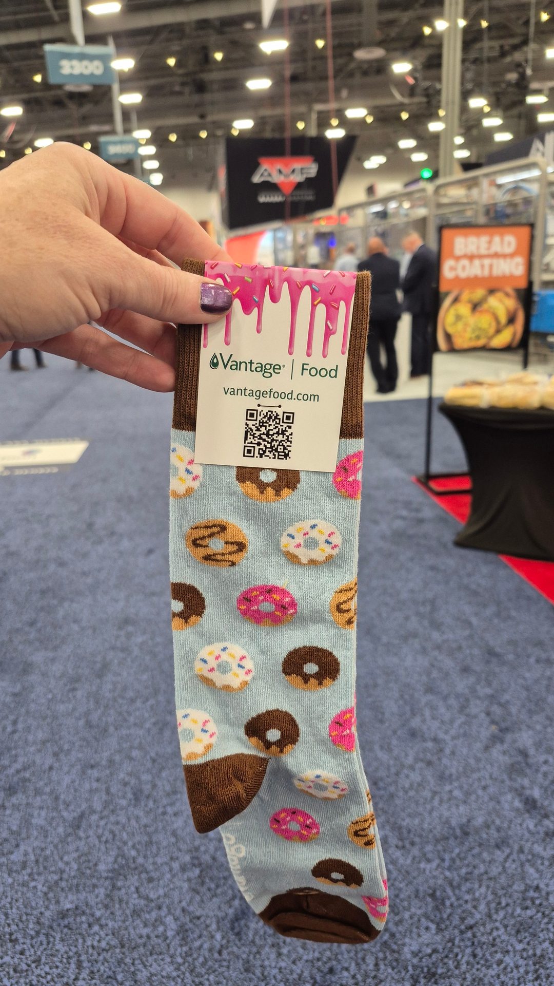 Hand holds a blue donut-patterned sock with a "Vantage | Food" tag at a trade show.