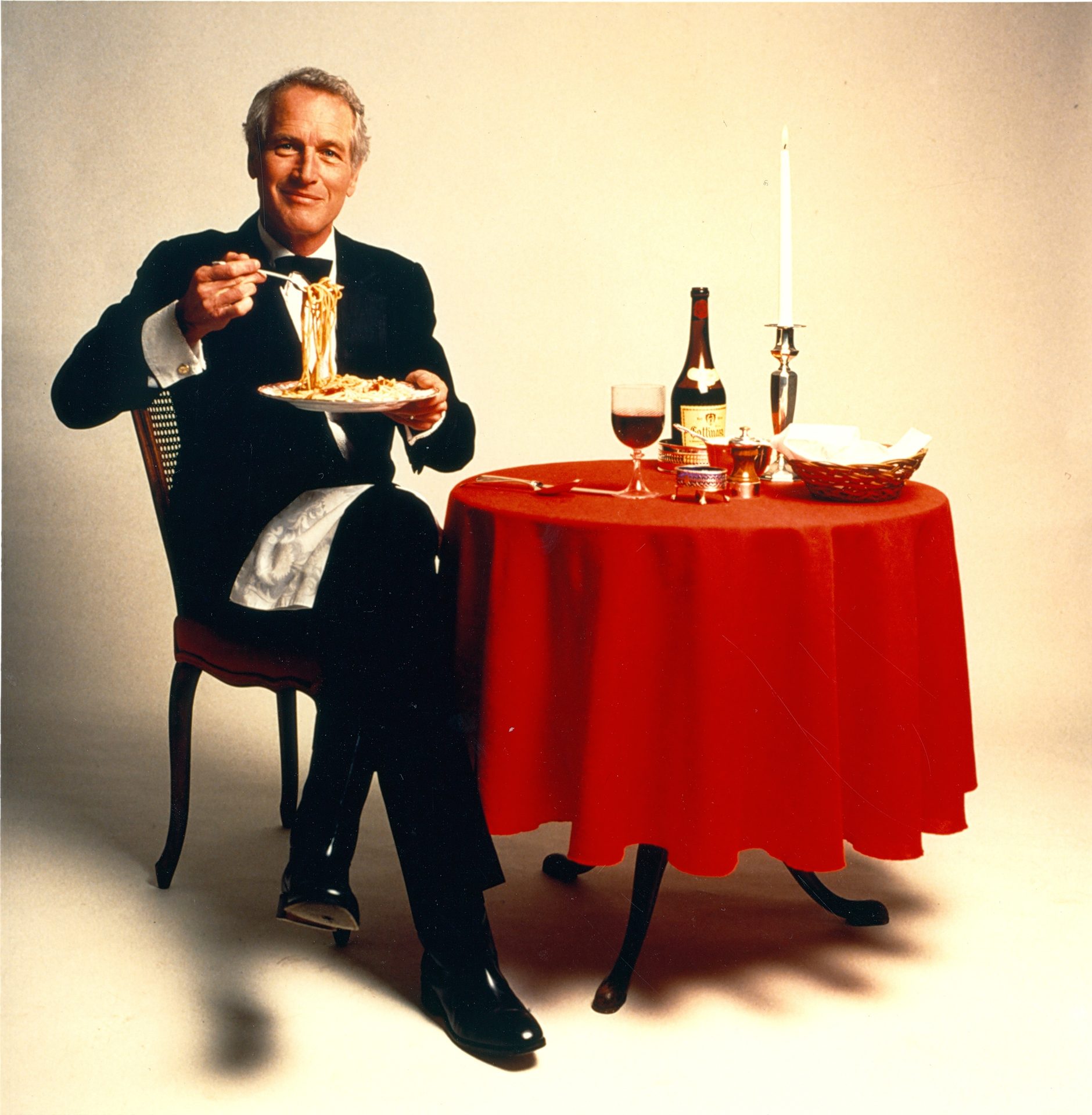 Paul Newman in a tuxedo, smiling while eating spaghetti at a table with wine and a candle.