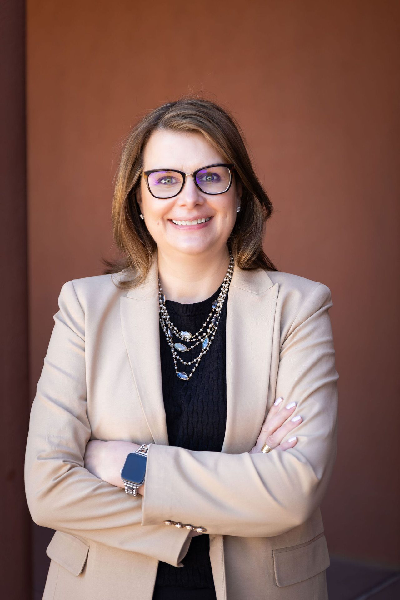 Professional portrait of a smiling woman in glasses, a beige blazer, and a layered necklace, arms crossed.