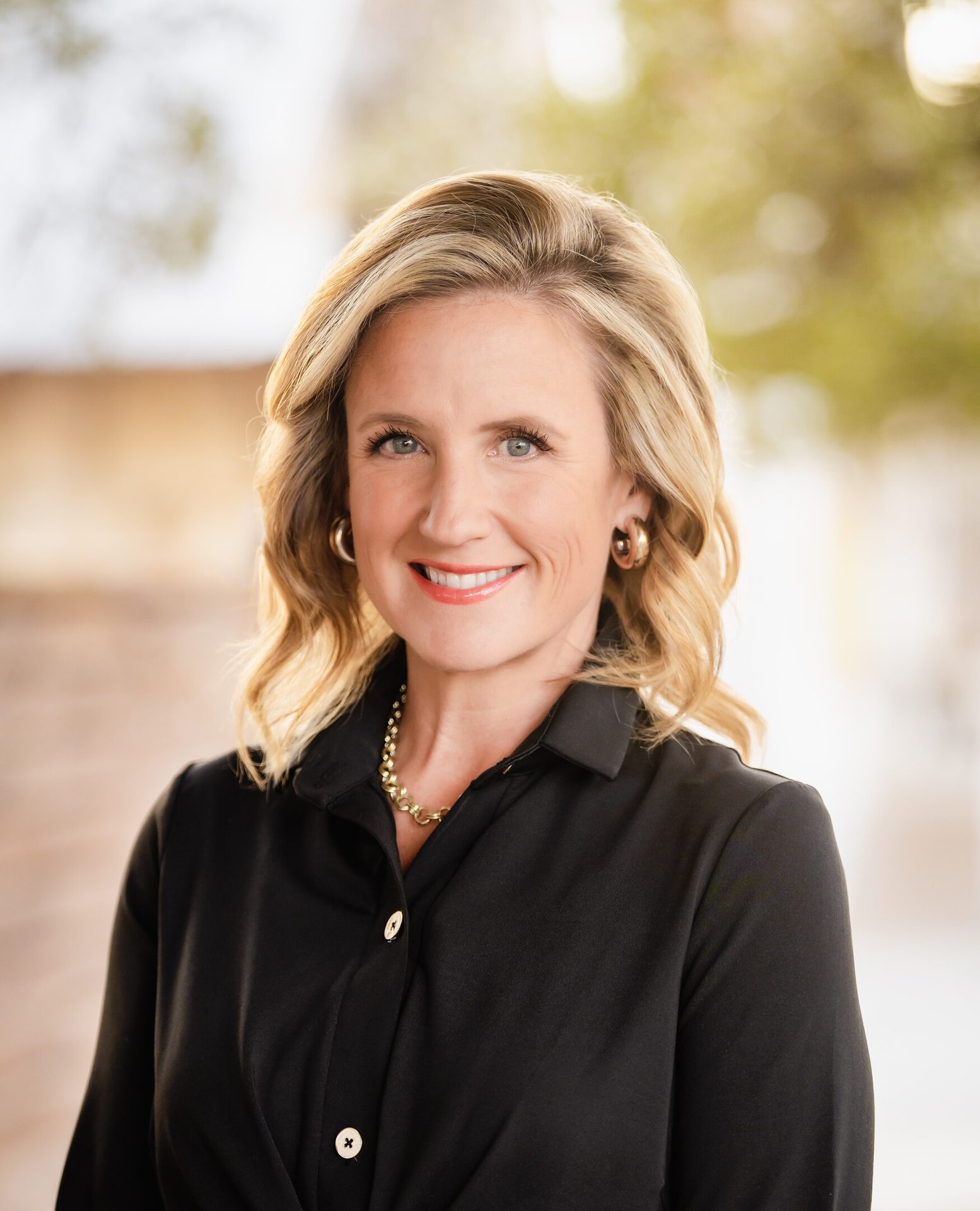 Professional headshot of a smiling blonde woman with blue eyes, wearing a black shirt and gold jewelry.