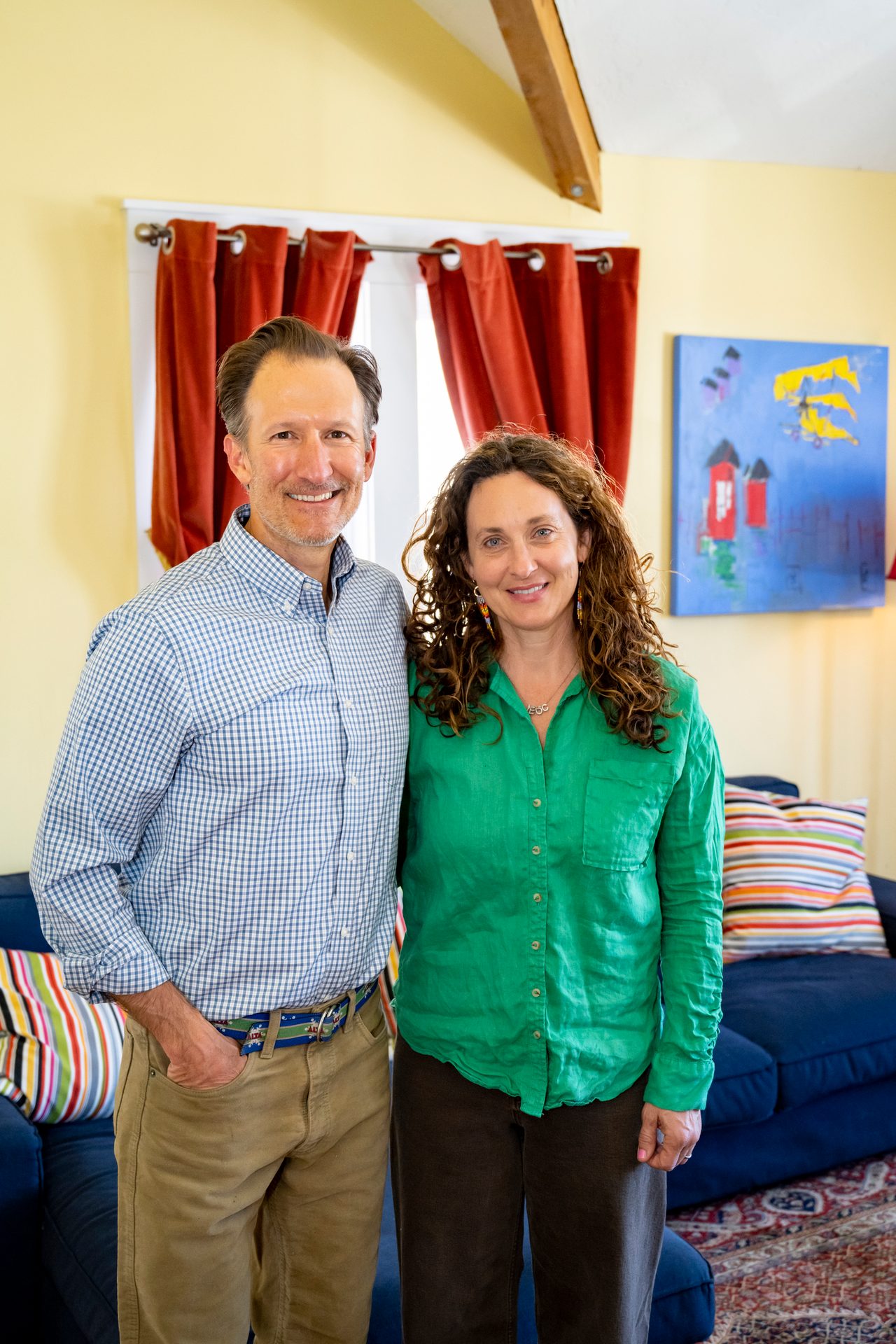 A man and woman stand side-by-side, smiling. He wears a plaid shirt, she a green one, indoors.
