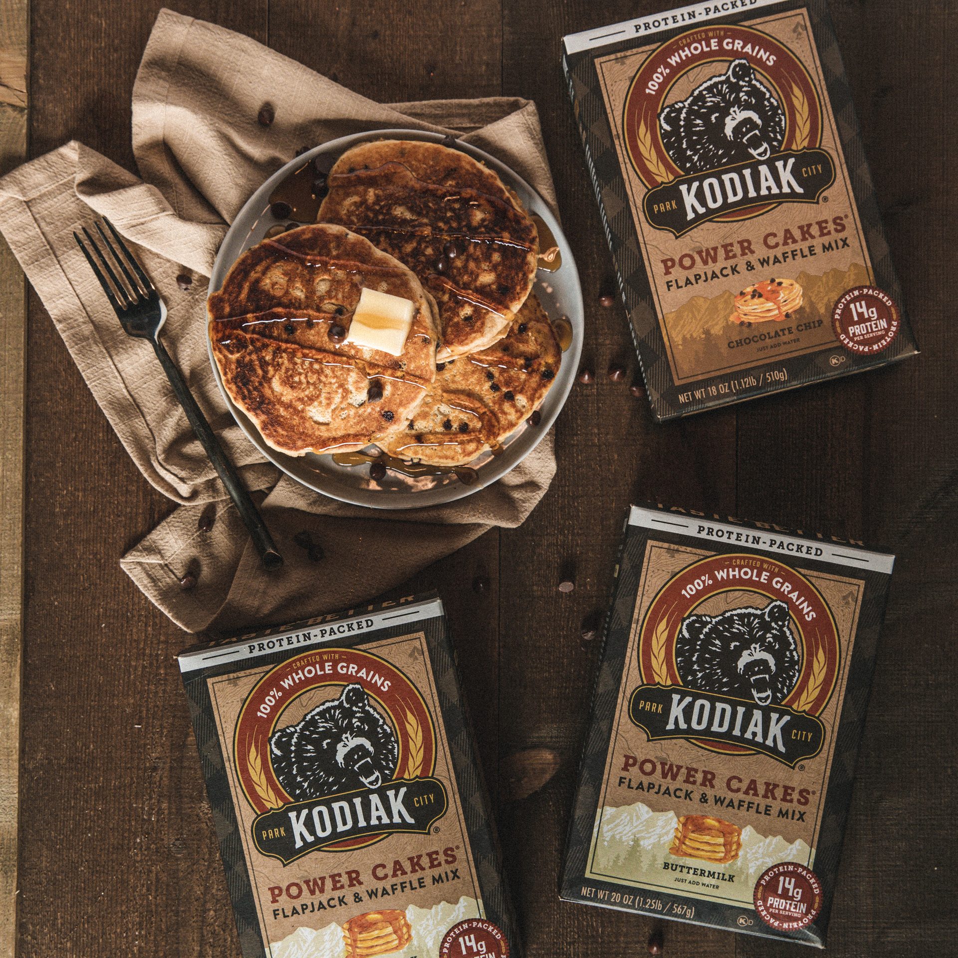 Kodiak Power Cakes Flapjack & Waffle Mix on a wooden table with finished pancakes on a brown napkin, with a fork