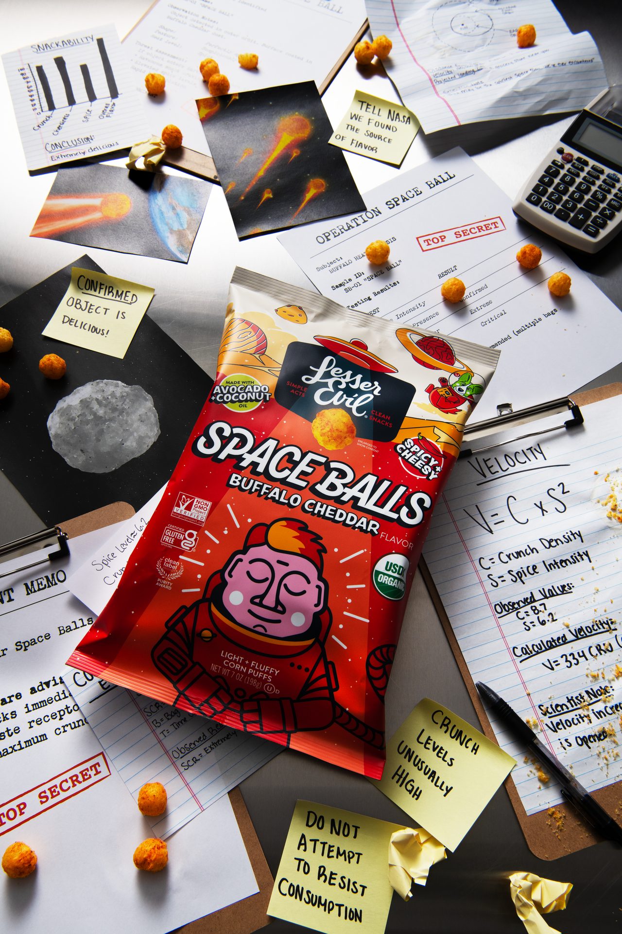A bag of 'Lesser Evil Space Balls' snacks surrounded by humorous space-themed 'scientific' documents and corn puffs.
