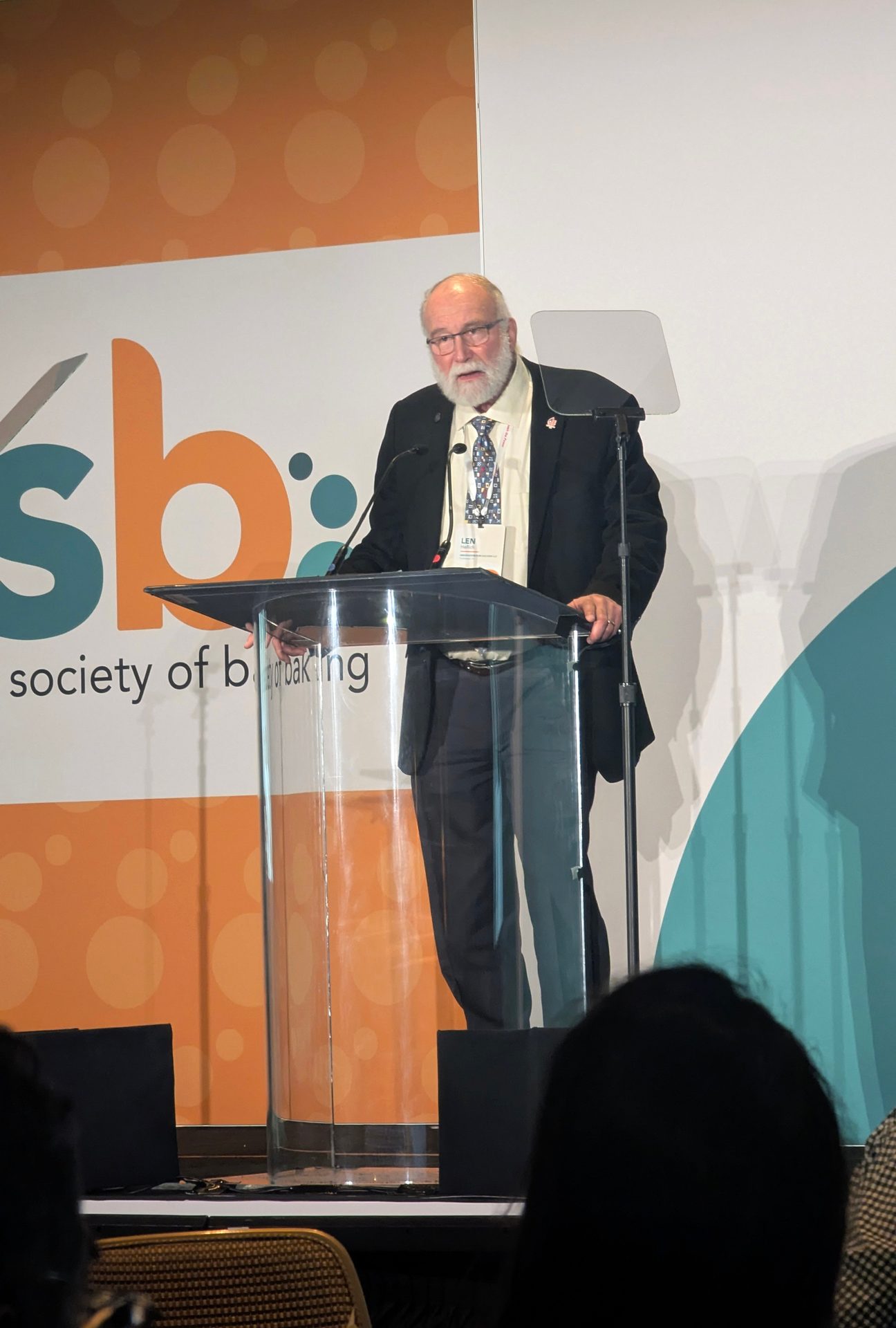Speaker in a suit with white beard and glasses at a conference podium.