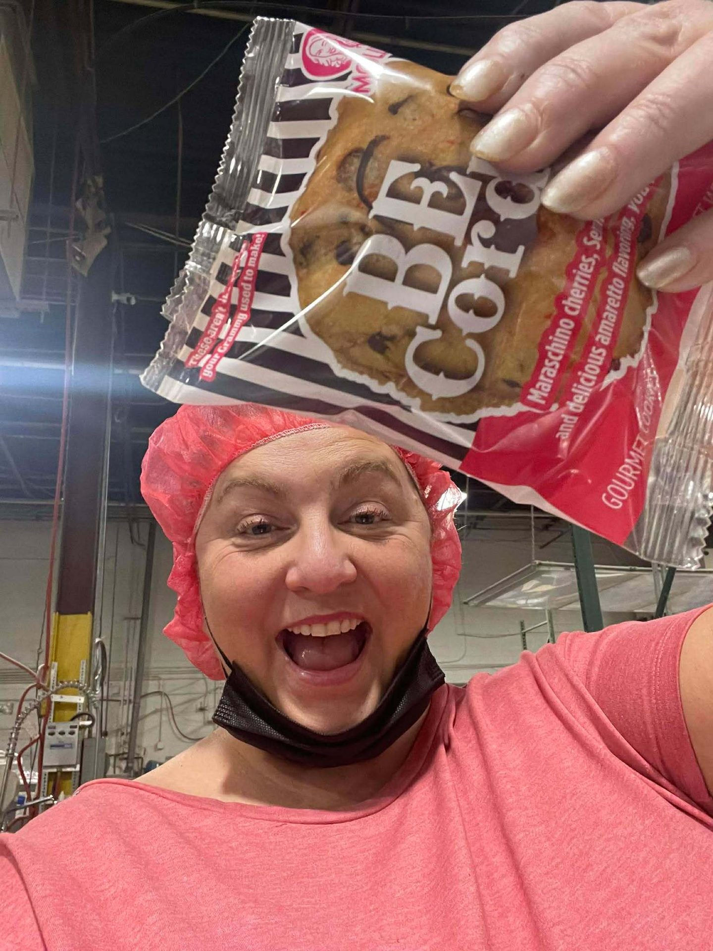 A woman in a hairnet and mask holds a "BE CORE" gourmet cookie package above her head, smiling excitedly.