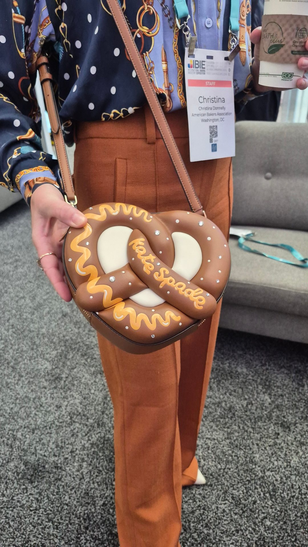 Person holding a pretzel-shaped Kate Spade purse, wearing a patterned shirt, orange pants, and an event badge.