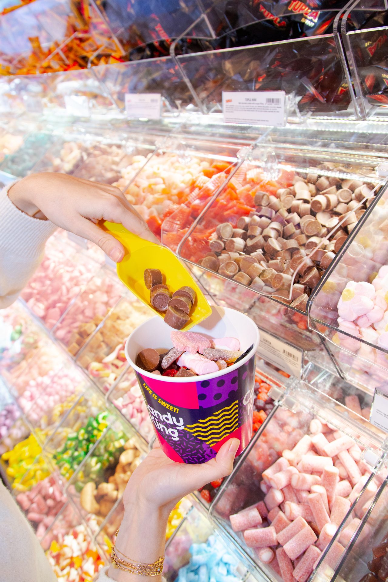 A person scoops chocolate candies from a bulk bin into a cup of mixed sweets at a candy store.