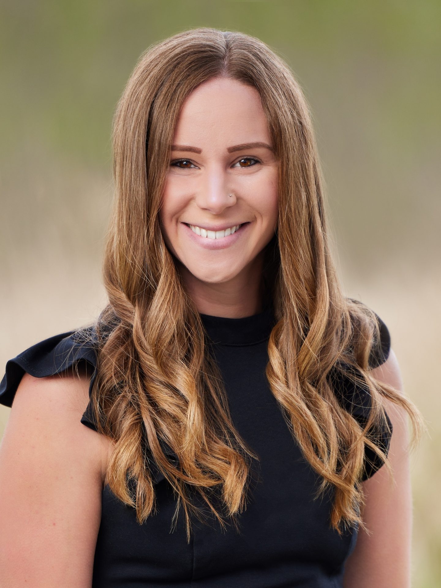 Smiling woman with long brown wavy hair, wearing a black top, blurred outdoor background.